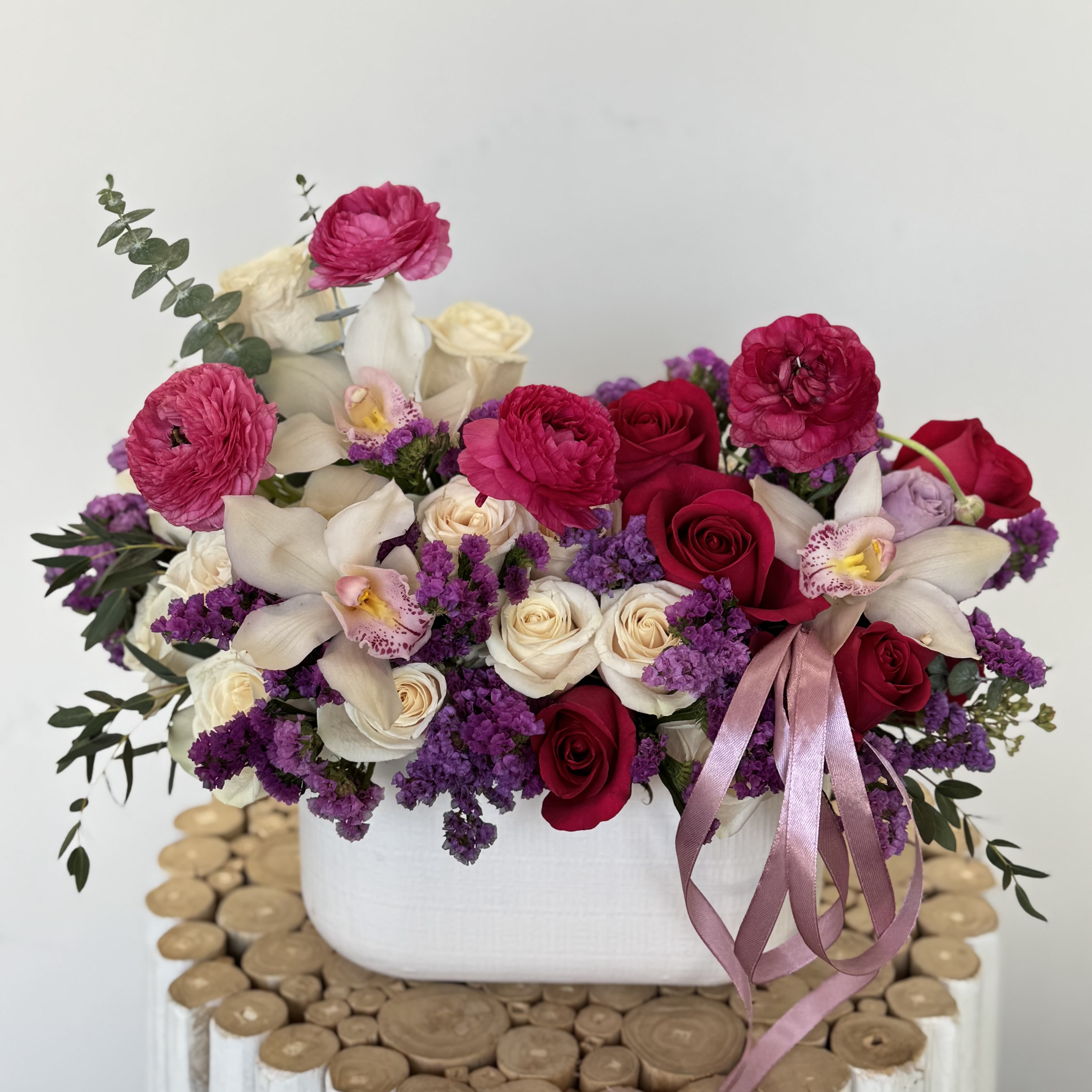 Garden Romance Arrangement - Fall in love with the Garden Romance Arrangement, a stunning mix of deep red roses, creamy orchids, ranunculus, and delicate purple fillers in a white ceramic planter. A sophisticated choice for anniversaries, engagements, or luxurious gifting.