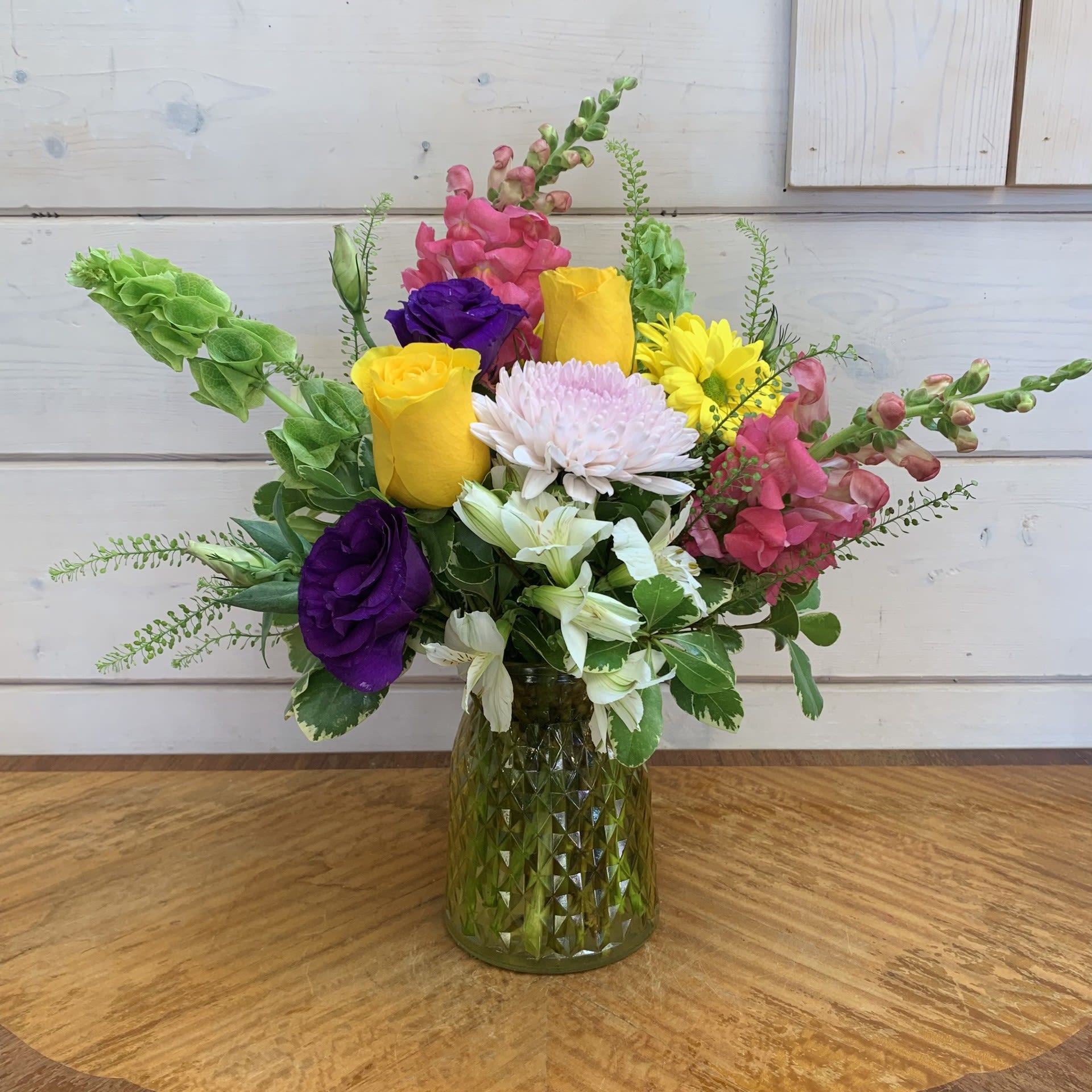 Bright Wishes - A vibrant arrangement of yellow roses, purple lisianthus, a cremon chrysanthemum, pink snapdragons, green bells of Ireland, and yellow daisies in a yellow tinted medium cut glass posie vase with white alstroemeria and pennycress to add a finishing touch of whimsy. Approximately 14&quot; tall and 12&quot; wide.