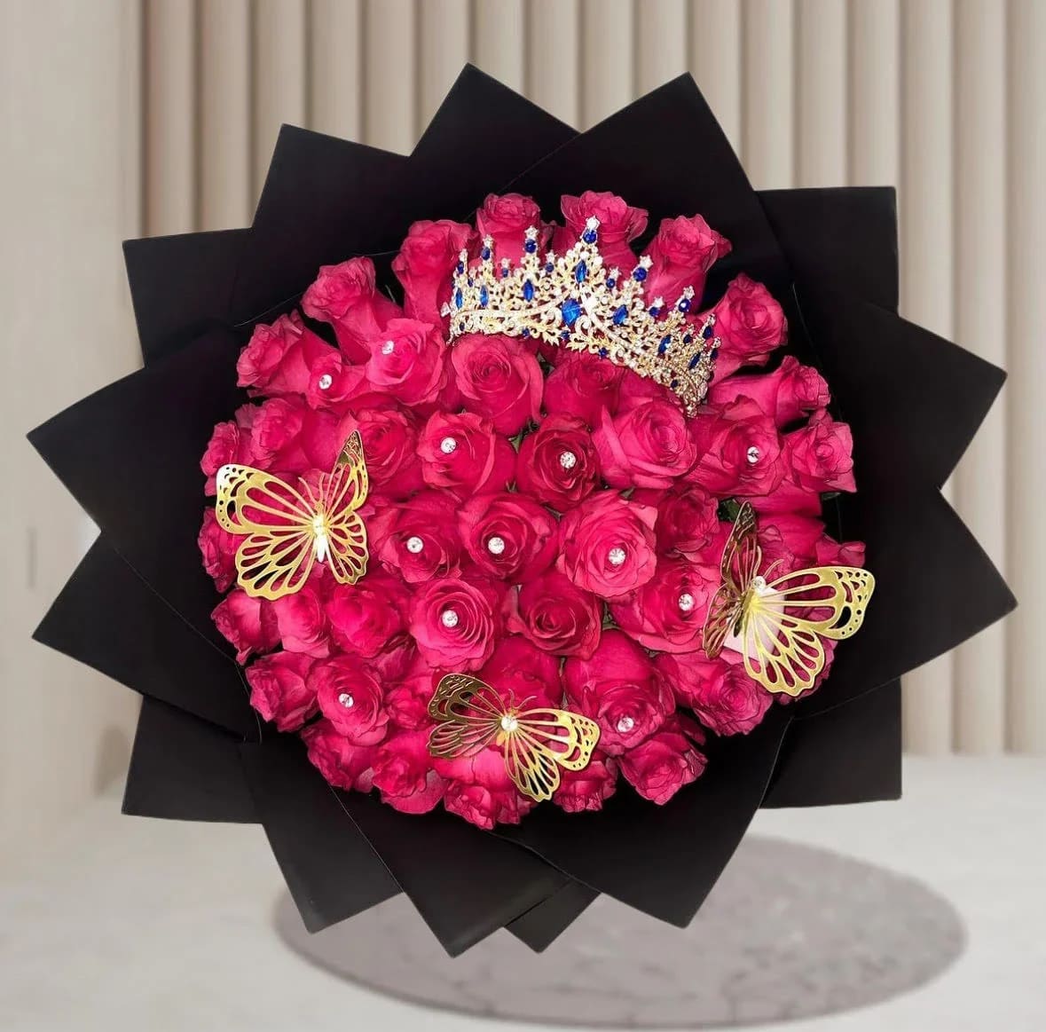 Queen Bouquet - A truly royal arrangement featuring vibrant hot pink roses adorned with delicate crystal accents. This bouquet is crowned with a dazzling jeweled tiara and decorated with elegant golden butterflies, symbolizing beauty and transformation. Wrapped in sleek black paper, it's the perfect gift for a queen on her birthday, quinceañera, or any special celebration.