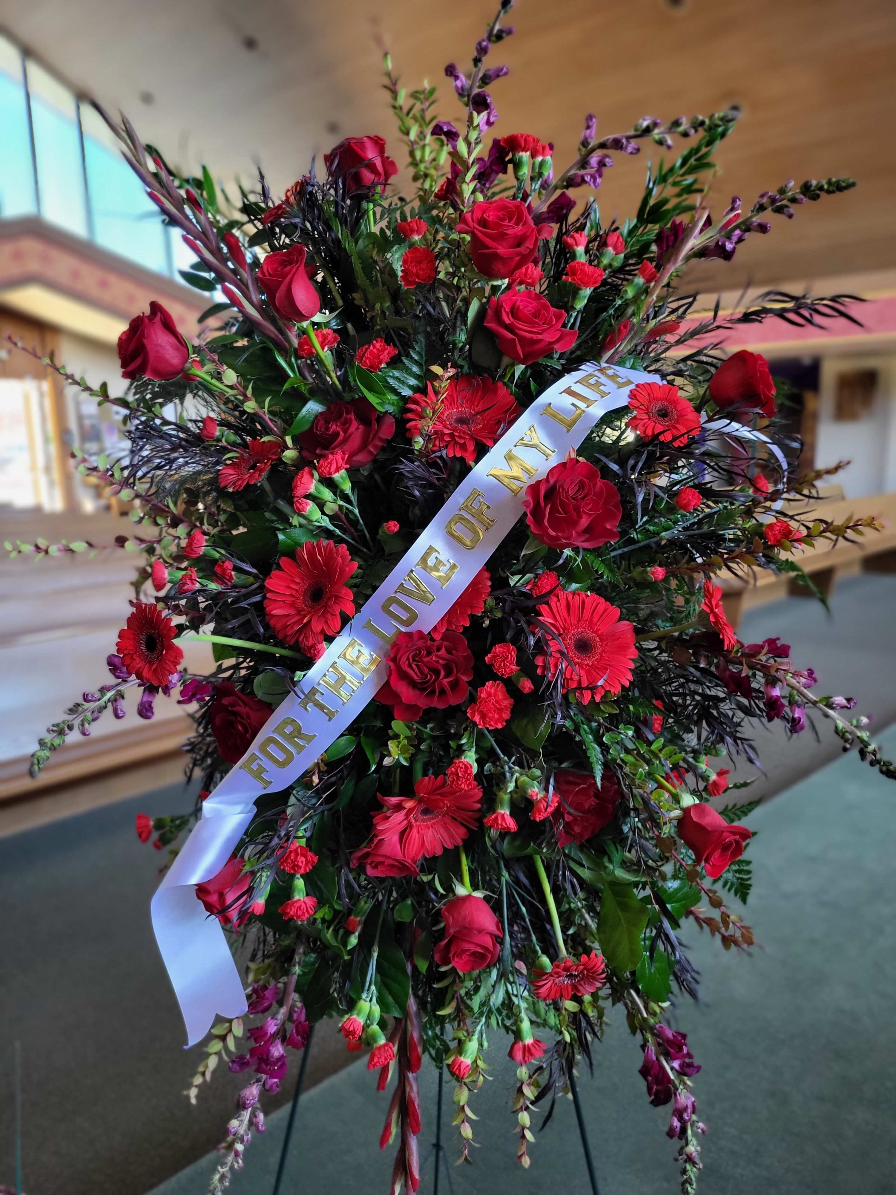 Large All Red Spray with Ribbon - Your Choice of Color  Order at least a week prior to the service and choose your color for this large spray tribute. We can downsize the scale or mix colors to bring the cost down if a large mono-color is out of budget. This item comes on a standing easel and includes an assortment of fresh flowers and greenery. Flower selection may vary.  Add a Custom Ribbon with Your Choice Sentiment   Shown here: Deep red, burgundy and crimson flowers and foliage.   Very large full spray on easel. Can be adjusted up or down in size and color change made with appropriate advance request.  Substiutions will be made especially when less time is given to acquire these flowers.