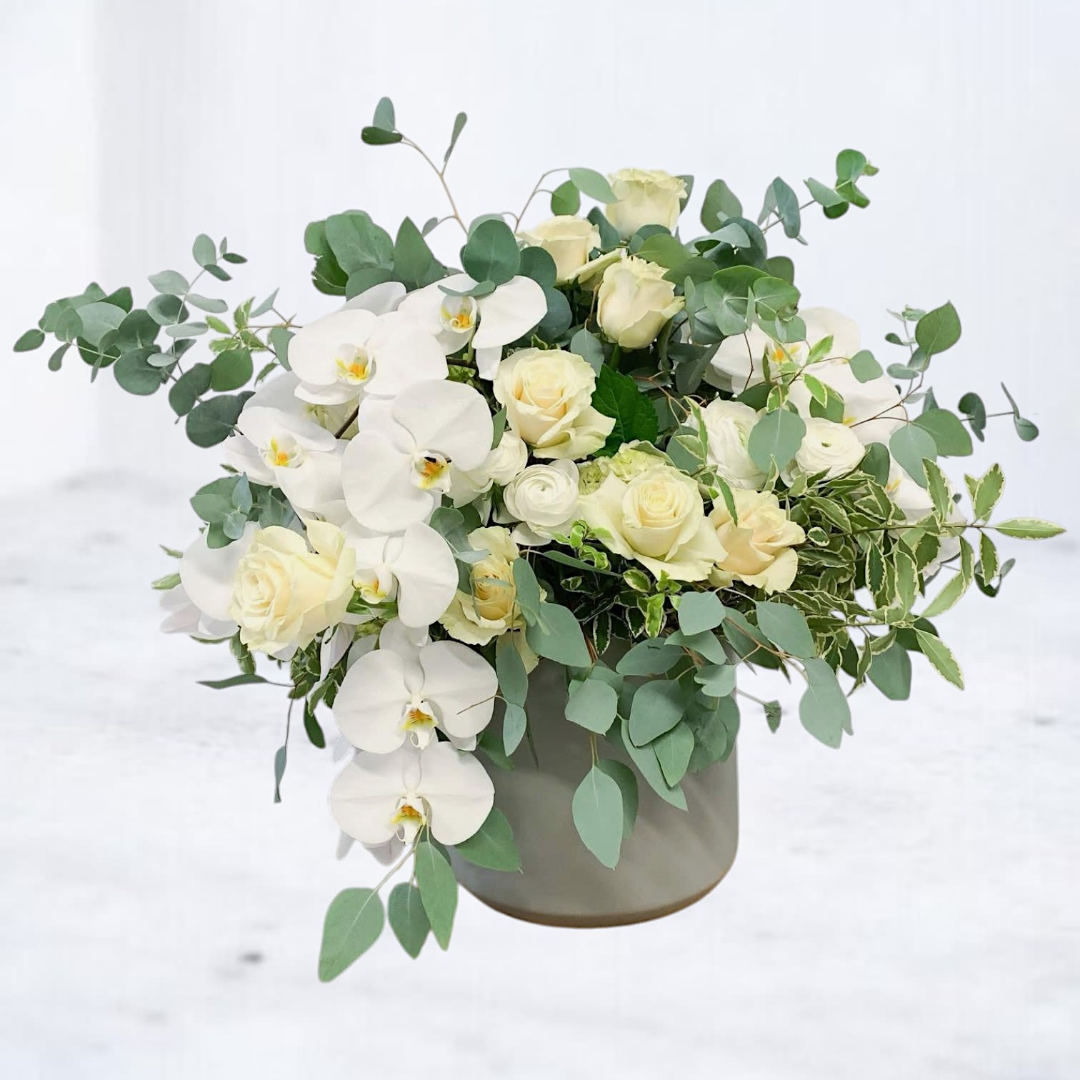 Bloom Chanson - A graceful symphony in soft white and green, Bloom Chanson is a floral composition that sings with elegance and poise. Featuring cascading Phalaenopsis orchids, creamy roses, ranunculus, and lush eucalyptus, this airy arrangement brings refined beauty to any space.  Designed in a modern matte vessel, this bouquet is ideal for sympathy flowers, upscale celebrations, or simply sending a note of sophisticated comfort. Its organic flow and timeless palette make it a standout choice for both heartfelt gestures and high-end decor. 