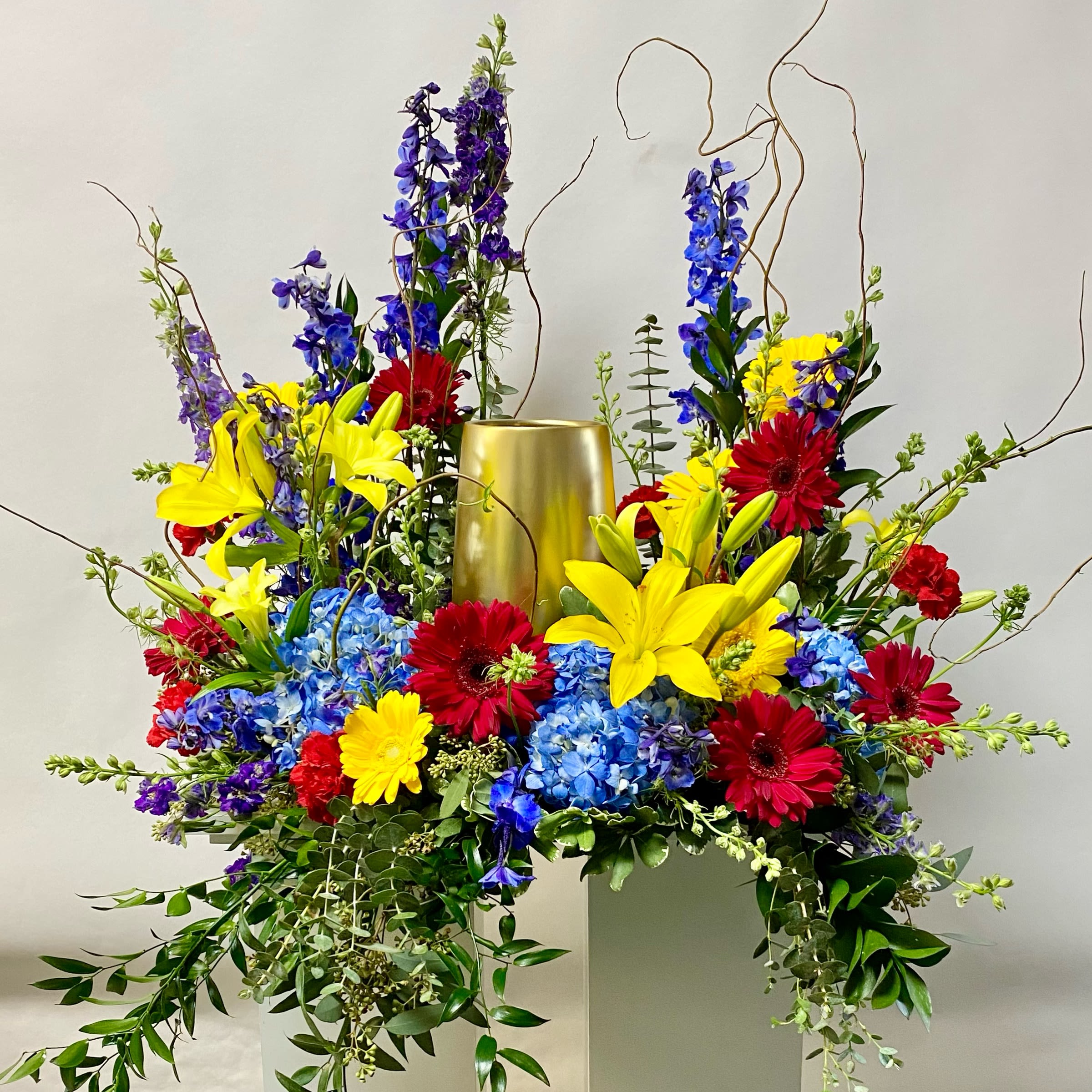 Sunny Garden Urn Wreath - This 18&quot; Urn Wreath Includes Purple Larkspur, Blue Delphinium, Red or Hot Pink Gerbera Daisies, Yellow Gerbera Daisies, Blue Hydrangea, Red Carnations and Yellow Lillies. Or as Similar as Possible. 