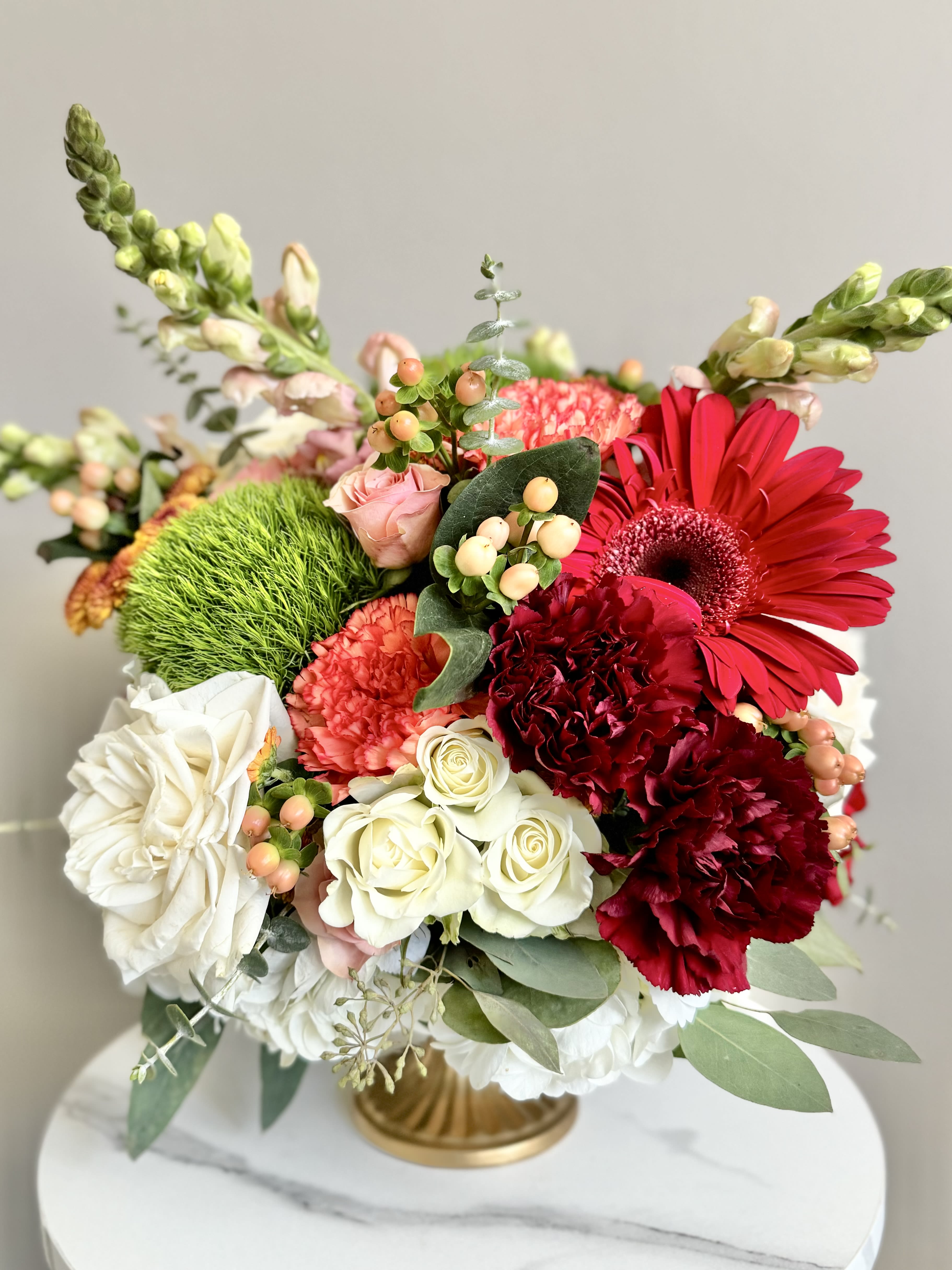 Table Centerpieces - A beautiful table centerpiece can transform an ordinary meal into an extraordinary experience. Imagine an elegant table setting, its warm tones contrasting with the soft glow of candlelight. At the center, a lush arrangement of flowers in soft, harmonious hues — pale blush carnations, red roses, and deep burgundy daisies — are nestled among sprigs of fresh greenery like eucalyptus and fern. The flowers are artfully placed in a low, elegant gold compote, allowing conversation to flow easily across the table. Imagine yourself being the best host that your guests will talk about for days, weeks, even years to come.   Flowers will be in season. Perfect for holidays, thanksgiving, birthdays, celebrations, and private parties. 