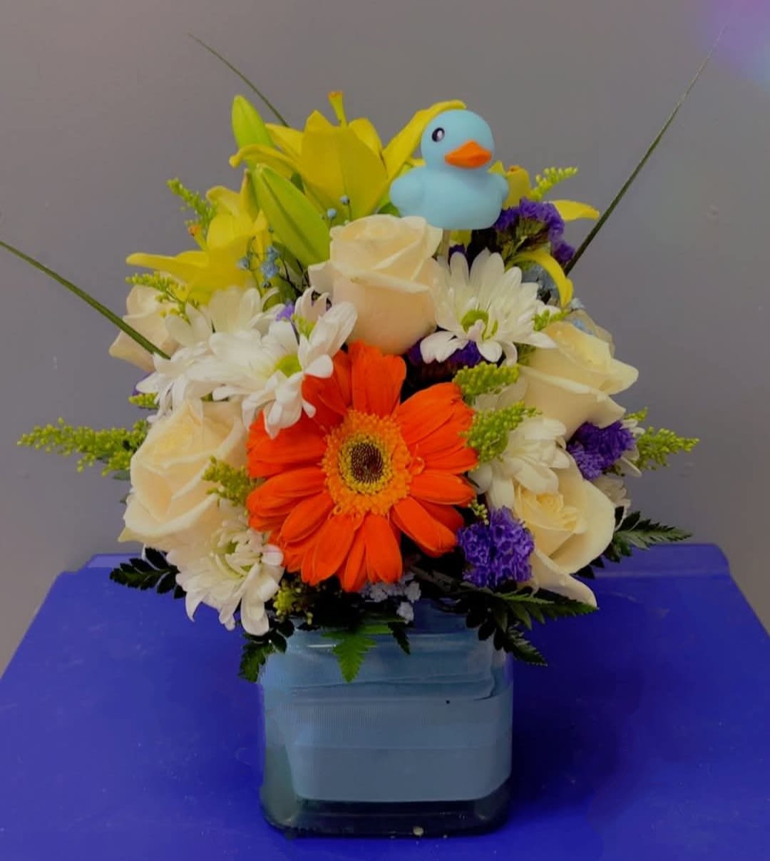 Oh, Boy! - Celebrate the arrival of a bouncing baby boy with our adorable Oh Boy! flower arrangement! Bursting with joy and vibrant color, this sweet design features an orange gerbera daisy, white roses, classic white daisies, bright yellow lilies, pops of purple statice, golden solidago, and various lush greens. All are artfully arranged in a baby blue cube vase and accented with a charming blue duck keepsake—perfect for the nursery. This cheerful bouquet is a delightful way to welcome the new little one and share in the happy news! 
