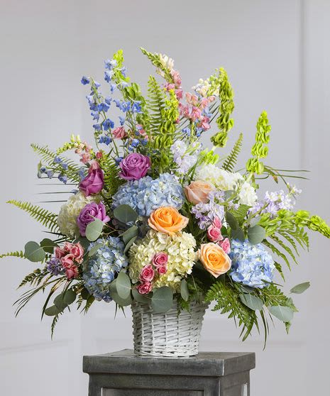 Garden Elegance Basket - A breathtaking floral arrangement bursting with the soft charm of a springtime garden. This elegant white woven basket features an artful blend of pastel-hued blooms including blue hydrangeas, peach and lavender roses, delicate snapdragons, and lush greenery. With its airy, organic feel and graceful height, this arrangement is ideal for expressing sympathy, celebrating milestones, or brightening any space with timeless beauty.