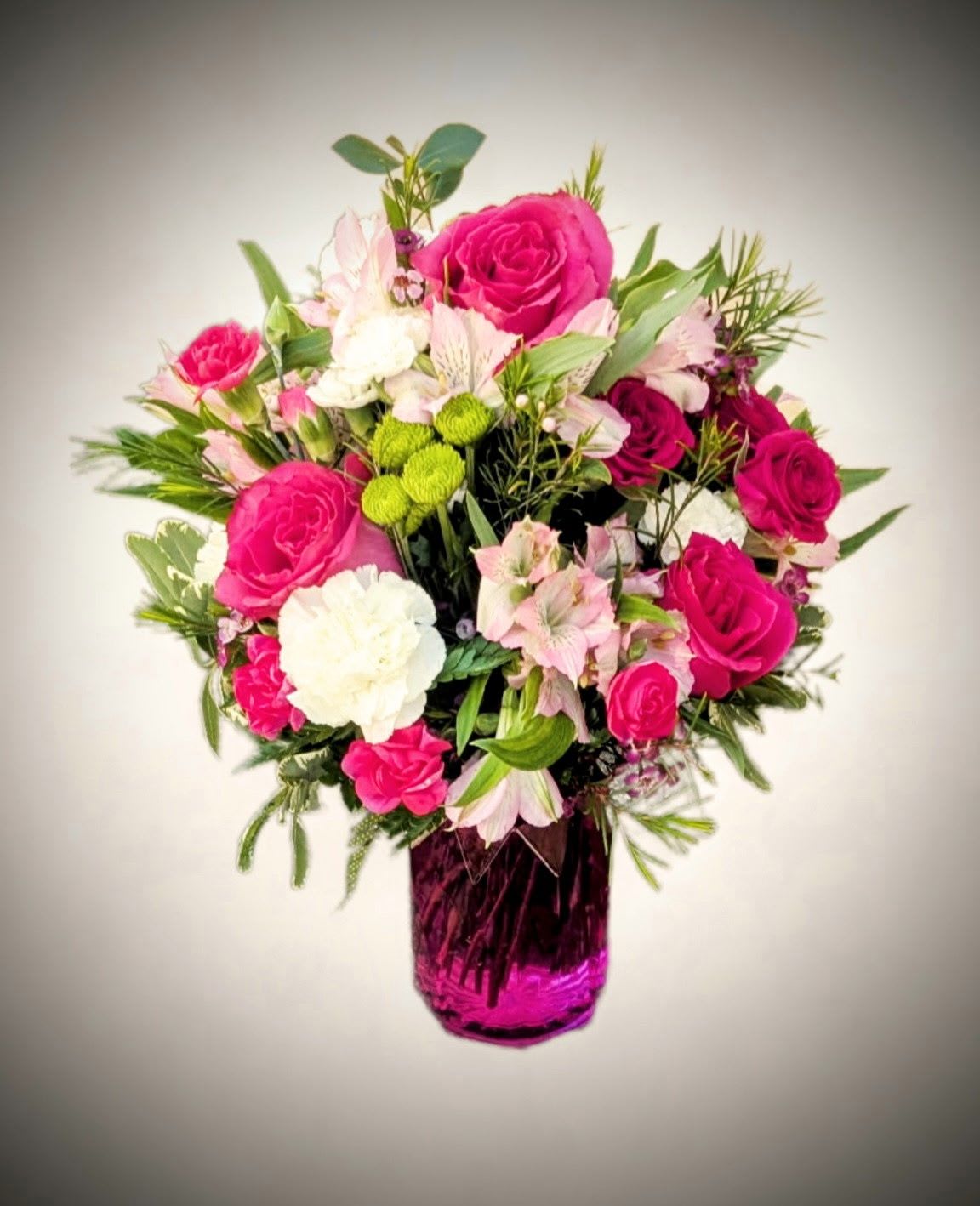Radiant Rose Garden Bouquet  - Brighten any space with the cheerful elegance of our Radiant Rose Garden Bouquet. This stunning arrangement features vibrant hot pink roses, soft blush alstroemeria, classic white carnations, and pops of green button mums, all artfully nestled among lush greenery and seasonal foliage. Presented in a striking fuchsia glass vase, this bouquet brings joy, color, and charm to any occasion—whether it's a birthday, anniversary, or simply to make someone's day.  Features:  Fresh hot pink roses and spray roses   Blush alstroemeria and white carnations  Accented with green button mums and mixed greenery  Delivered in a deep pink glass vase  Perfect for birthdays, celebrations, or everyday cheer   Approximate Dimensions: 14&quot; H x 12&quot; W   