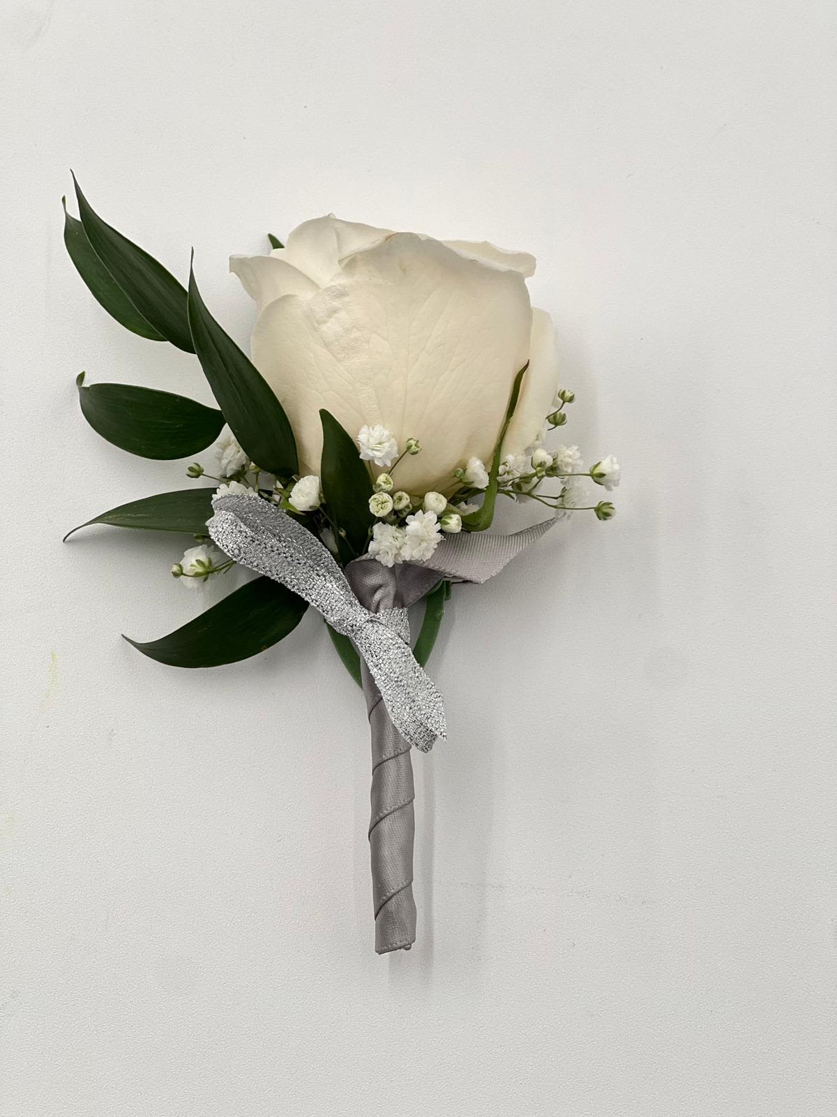 Boutonnière 4 Boutonnière 4 - A timeless boutonnière featuring a single, pristine white rose, delicately accented with baby's breath and lush greenery. Wrapped in a sleek silver ribbon and finished with a touch of sparkle, this elegant floral accessory adds a classic and refined touch to any formal occasion, perfect for weddings, proms, or special celebrations.