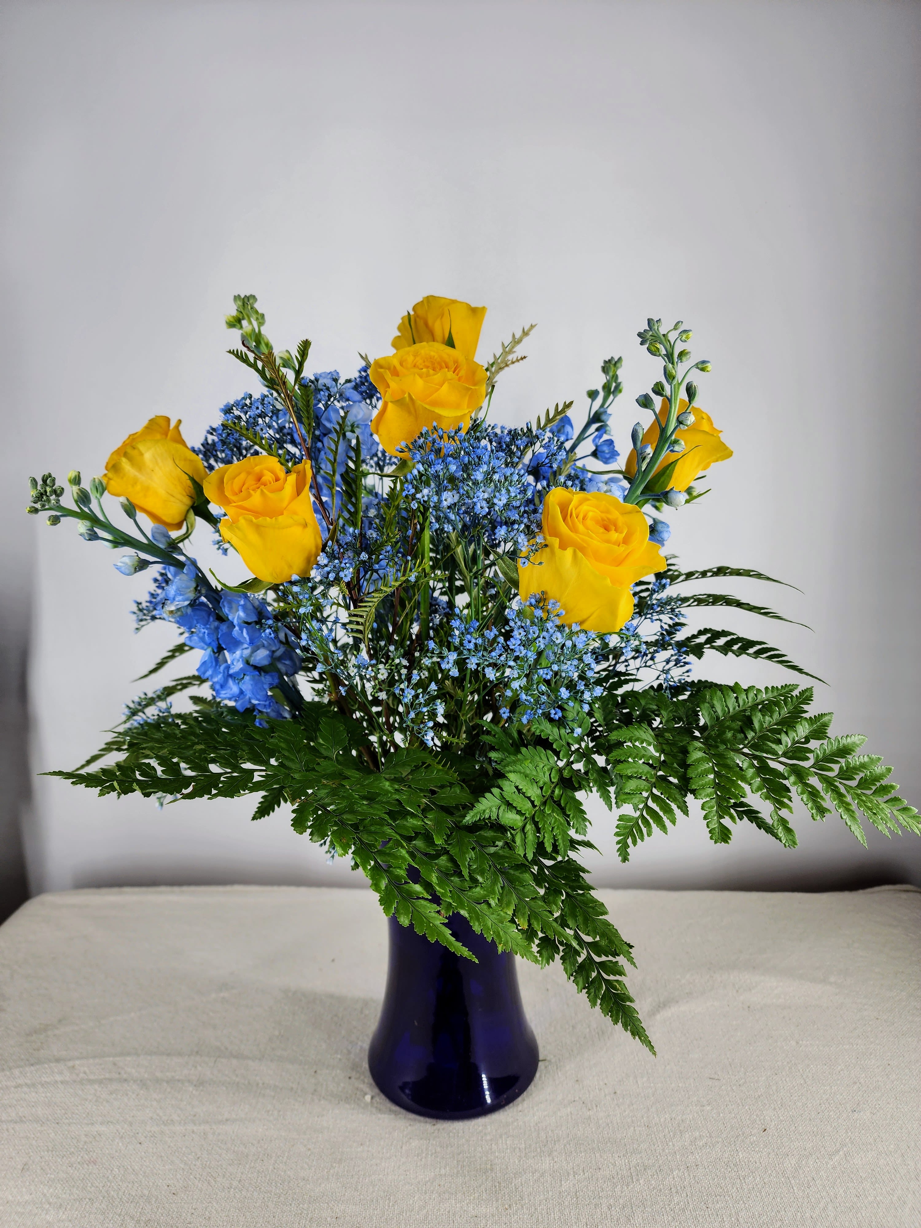 Swedish Style - Yellow roses engulfed in blue stock, blue baby's breath a assorted greens in a blue vase.
