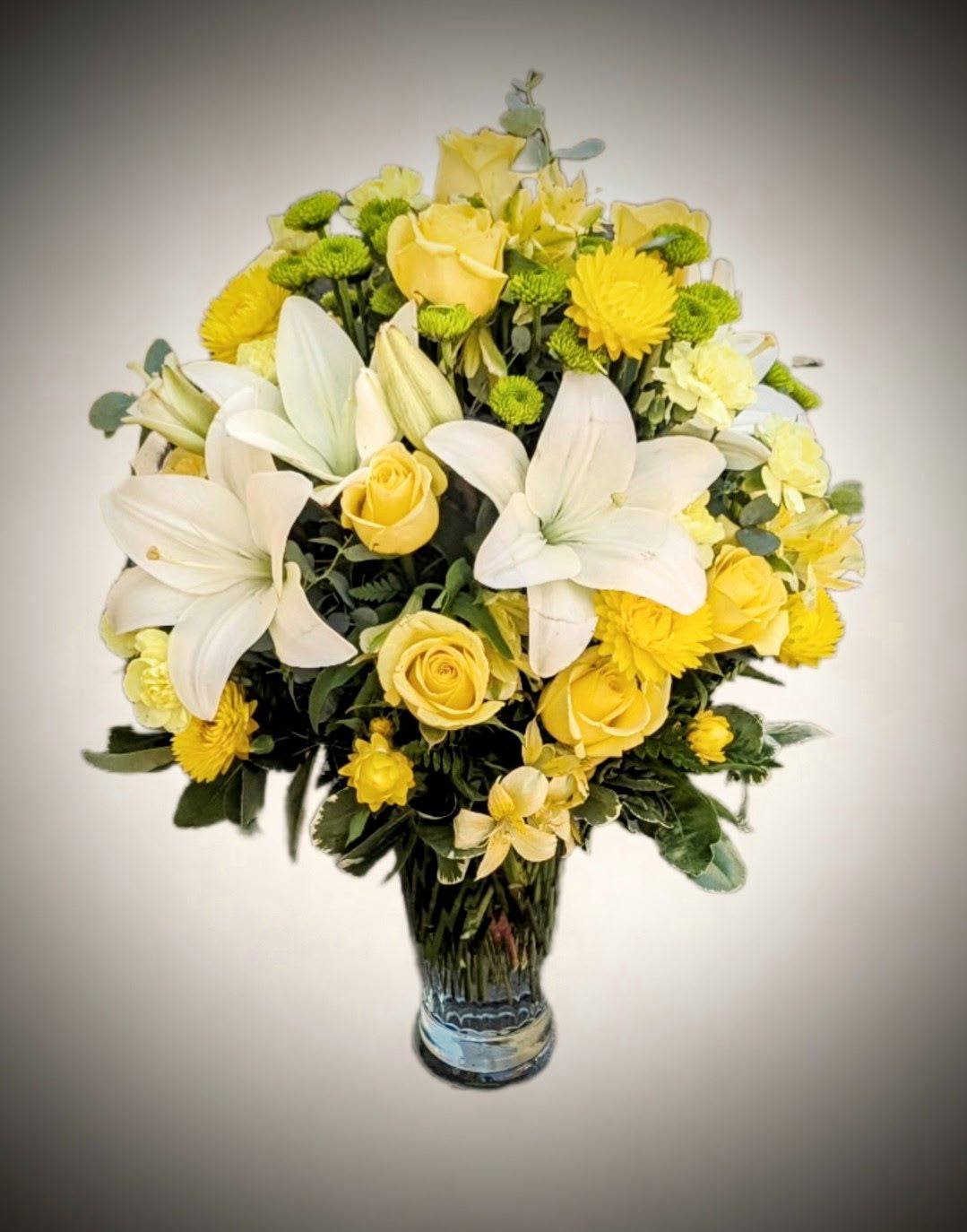 Golden Radiance Bouquet  - Elevate any occasion with the timeless beauty of our Golden Radiance Vase Arrangement. This stunning bouquet showcases an exquisite mix of creamy white lilies, soft yellow roses, golden chrysanthemums, vibrant green button poms, and delicate filler blooms — all thoughtfully arranged in a classic clear glass vase.  A symbol of joy, light, and warmth, this radiant bouquet is ideal for celebrations, sympathy gestures, or simply brightening someone's day. Its soft yet uplifting color palette brings a serene elegance to any room.  Features:  Fresh-cut premium flowers including lilies, roses, mums, and poms  Arranged in a tall, stylish clear glass vase  Perfect for birthdays, anniversaries, sympathy, or get-well sentiments   Let love and light blossom beautifully with this graceful floral display