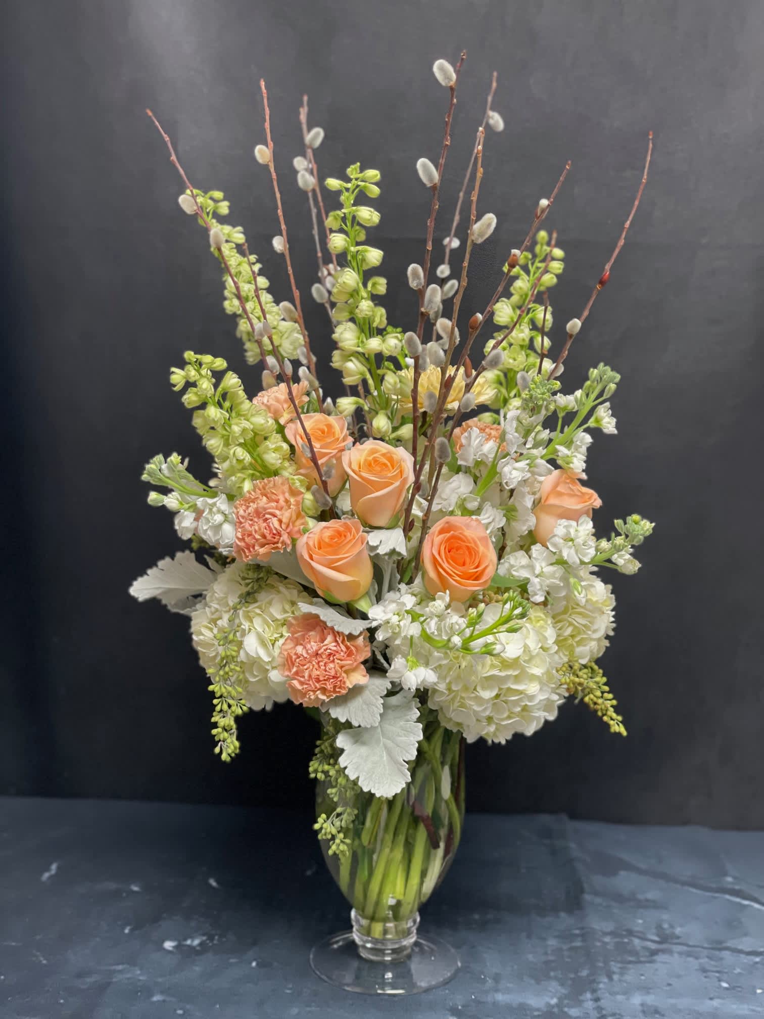Peach Blossom Bouquet - White Hydrangeas, Peach Roses, Peach Carnations, White Stocks, White Delphiniums, Peach Mums, Dusty Millers, Pussy Willows and Seeded eucalyptus in a clear glass vase.  The arrangement is all around.