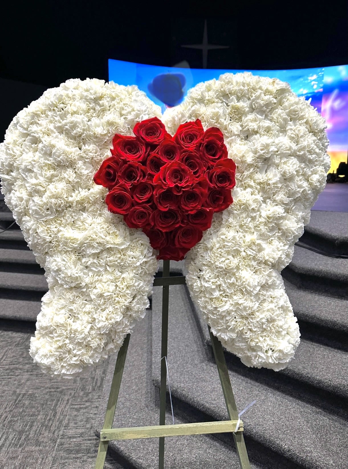 Angel Wings Floral Tribute with Heart - This striking floral arrangement is designed in the shape of angel wings, symbolizing peace, purity, and the ascension of a loved one. Crafted with dense clusters of white carnations, the wings encircle a vibrant red heart formed by a collection of fresh red roses, representing deep love and eternal remembrance. Displayed on a standing easel, this elegant tribute is often used for memorials or funeral services to honor and celebrate a departed soul, evoking both heavenly protection and heartfelt affection.  ********10 DAY ADVANCE PURCHASE*********** ***CUSTOMIZATION OF HEART COLOR AVAILABLE***