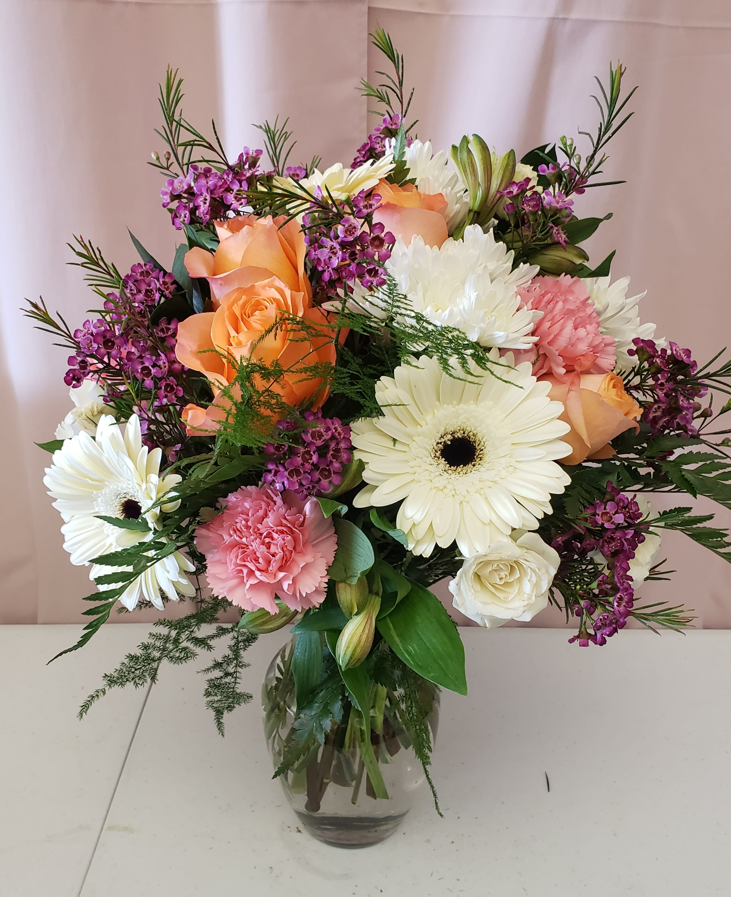 A Mother's Love - Bright and happy Mother's Day arrangement sure to make that special Mom smile!  Vase arrangement with white gerbera daisies, peach roses, white mums. pink carnations, and elegant purple waxflower. 