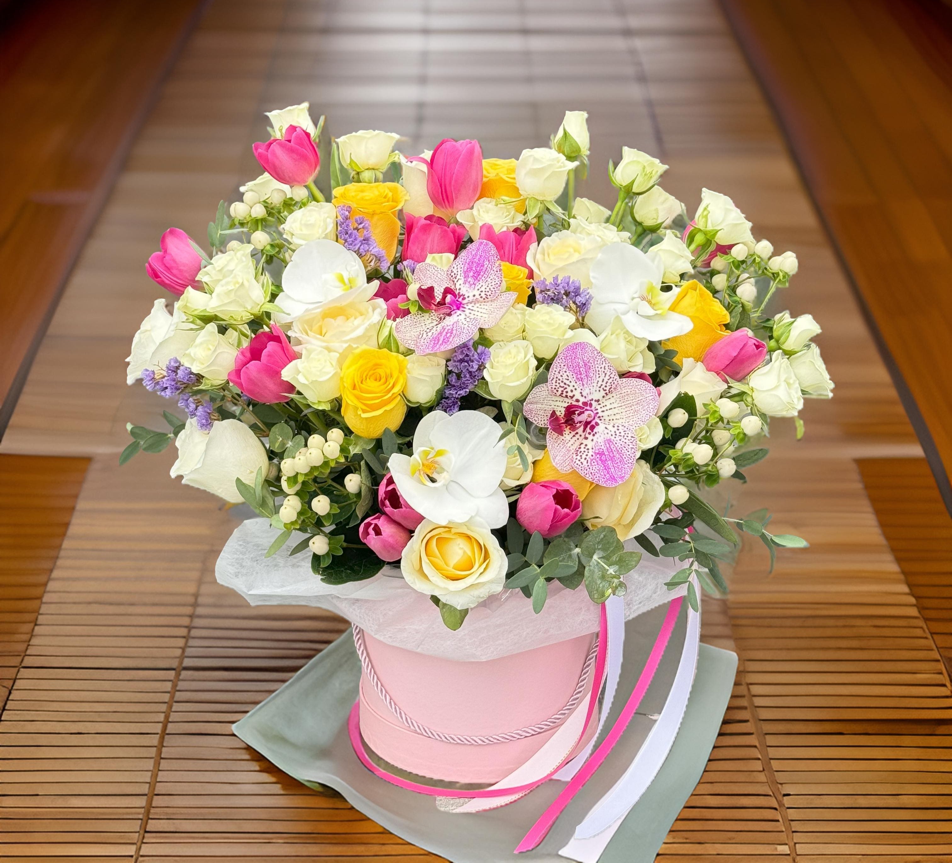 Sunshine Bloom Box - This vibrant flower box is a bold celebration of color and elegance. Expertly arranged with fresh yellow roses, white spray roses, pink tulips, and delicate orchids, it brings together classic blooms and exotic details in one luxurious composition. The bright and cheerful palette makes it a perfect gift for birthdays, baby showers, graduations, or as a stylish centerpiece for a brunch, bridal shower, or event.  Presented in a high-quality round hatbox, this floral arrangement adds a modern twist to traditional flower gifting. The soft pink box creates a beautiful contrast to the vivid colors, making it a standout piece for any space.  Ideal for flower delivery in Los Angeles, this arrangement suits any occasion where beauty, joy, and elegance are appreciated. Whether you're sending congratulations, appreciation, or a surprise gift, this flower box is the perfect expression of style and positivity.