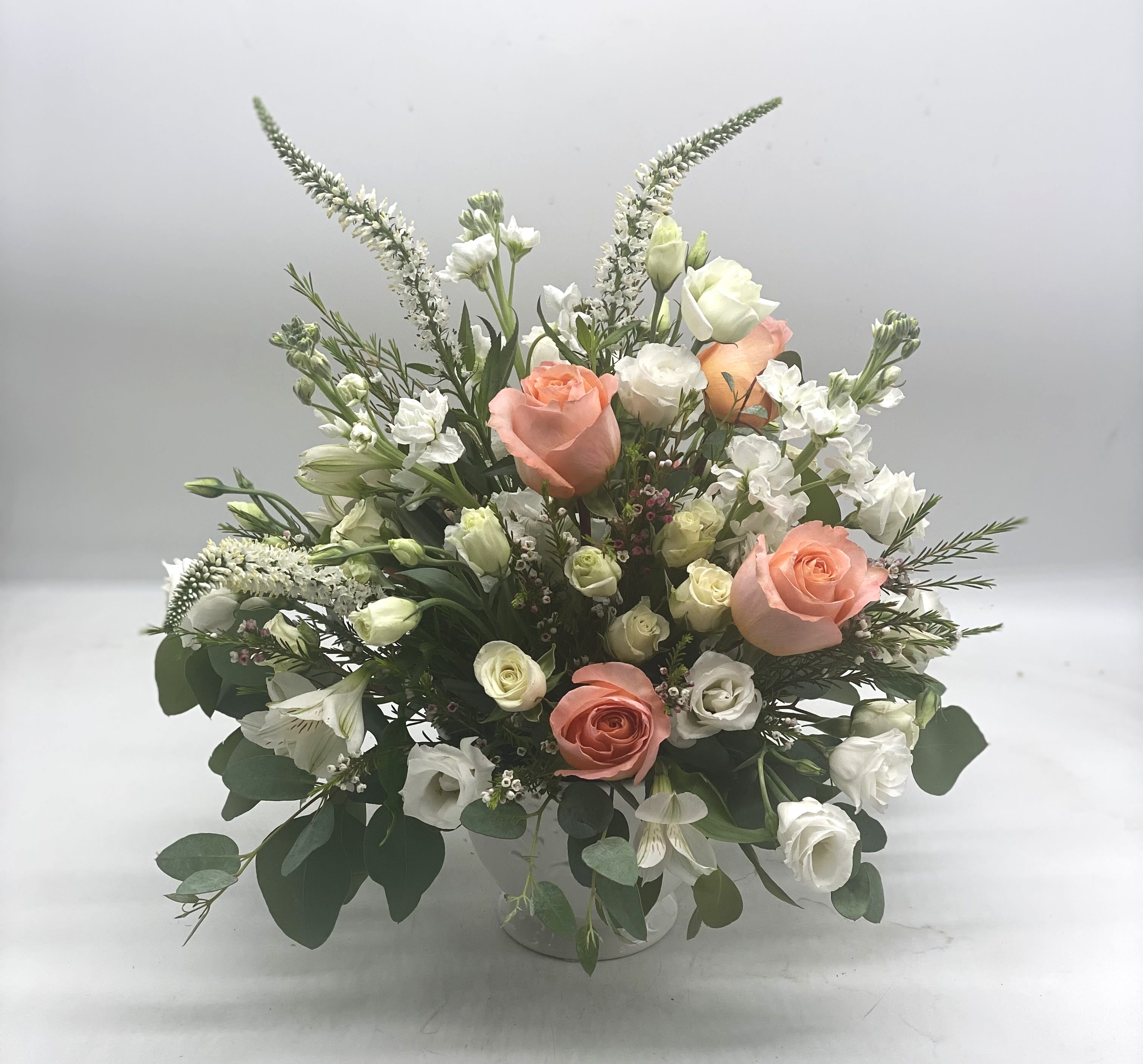 Coral Whisper - This arrangement features soft coral roses as the focal point, complimented by cream white lisianthus, spray roses, stock, veronica and sprigs of wax flower and eucalyptus, designed in a white ceramic compote bowl. Great for anniversaries, sympathy or simply to bring a touch of peace into any space!