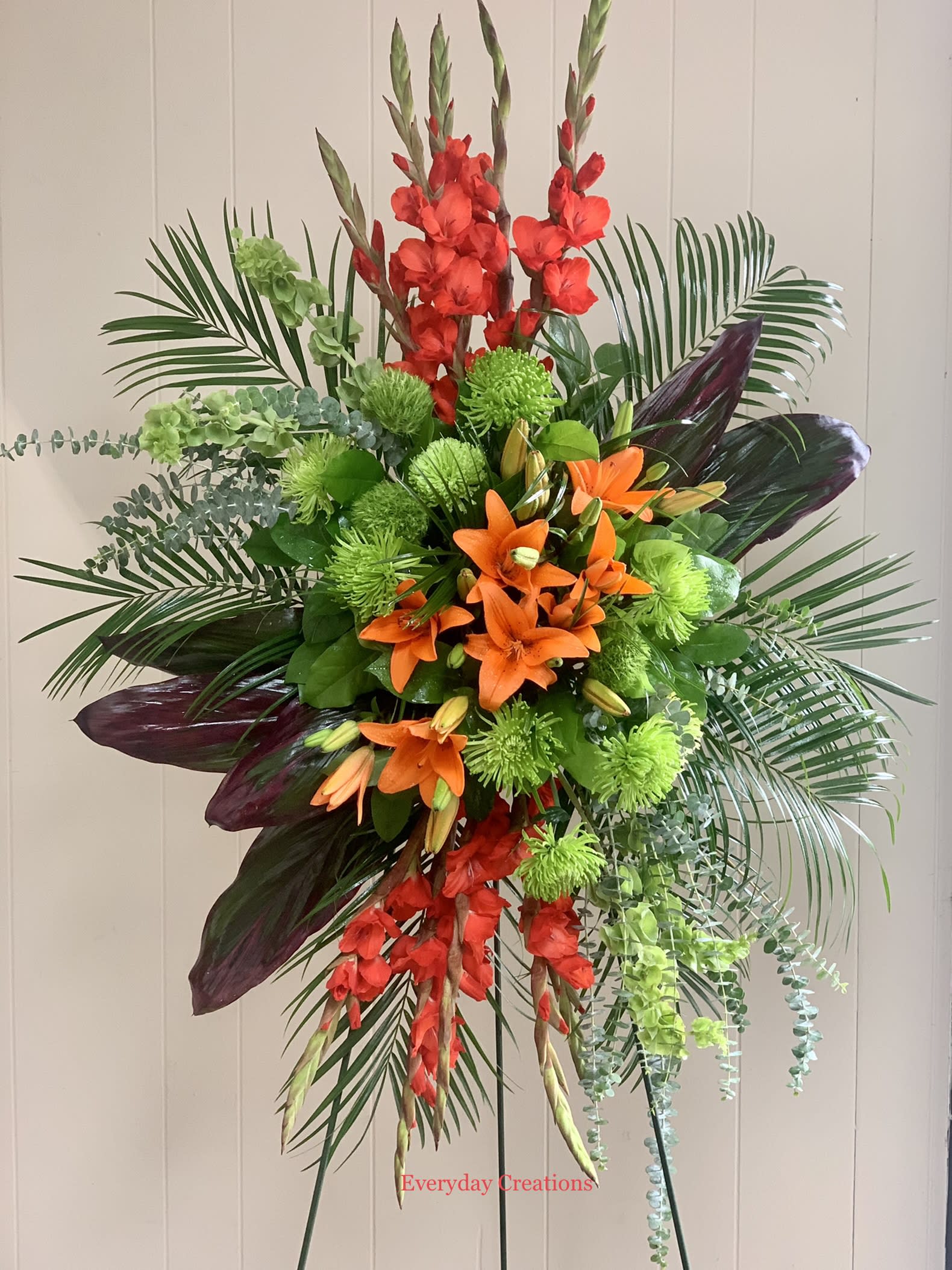 SYM16 - Sunrise Spray SYM16 - Sunrise Spray - The Sunrise Spray standing arrangement is a beautiful and unique way to pay tribute to your loved one. this tropical style arrangement features a stunning combination of orange lilies, red gladioli, green carnations, peach hypericum, emerald palm and red ti leaves, creating a vibrant and energetic display. The Sunrise Spray is delivered on a sturdy easel, making it easy to display at a funeral service. It is a beautiful and meaningful way to honor your loved one and celebraate their life.