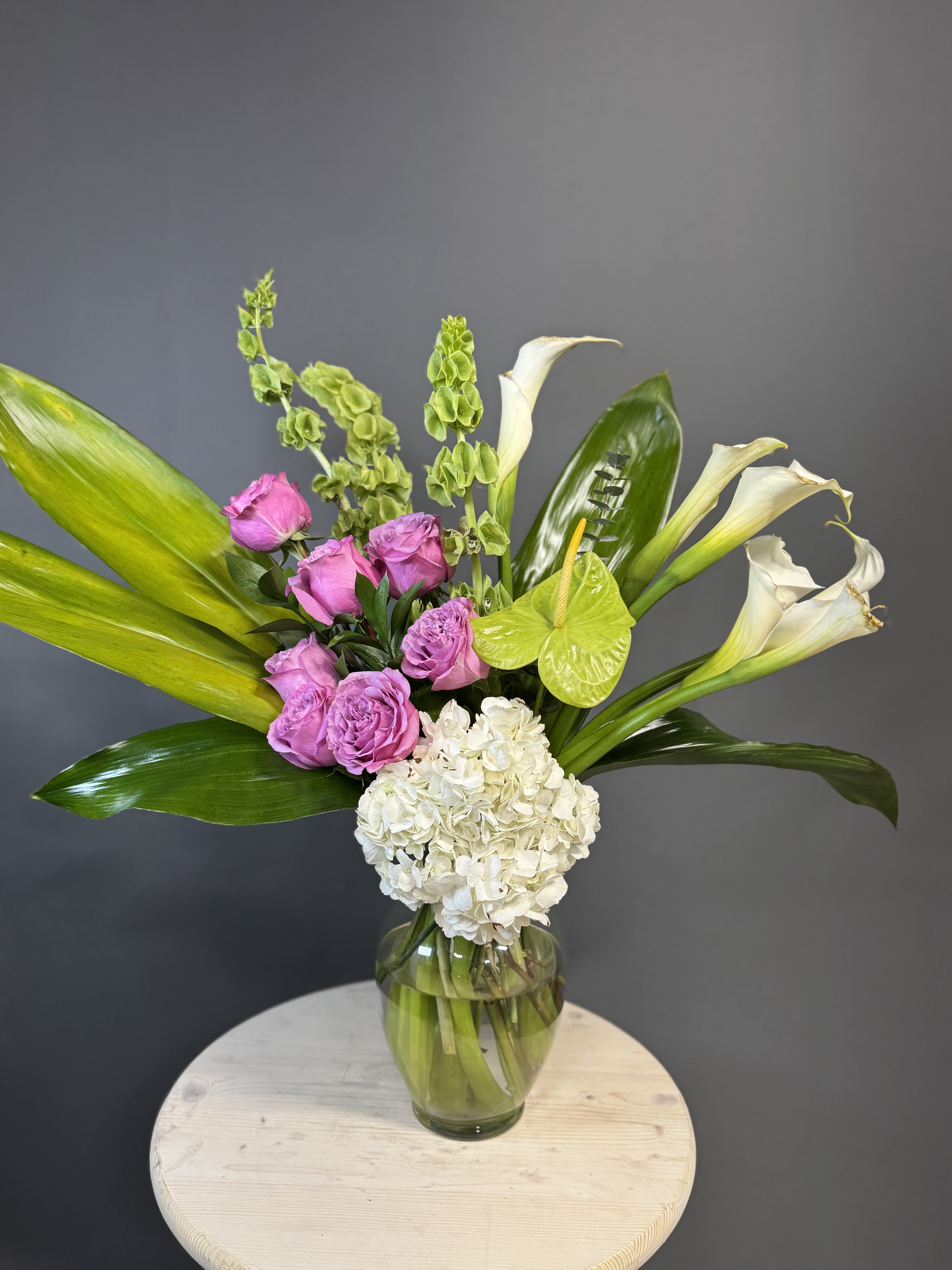 Kyoto Charm Bouquet - Introducing our &quot;Kyoto Charm Bouquet&quot; - a delightful mix of white hydrangeas, calla lilies, green bells of Ireland, Anthuriums, and purple roses in a clear vase. This arrangement captures the essence of Kyoto’s charm with a playful blend of colors and textures. Perfect for bringing a touch of whimsical beauty into any space.