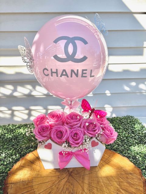 CC Balloon and Bag - A wooden box filled with roses is a timeless and elegant floral arrangement. The contrast between the rustic charm of the wooden box and the delicate beauty of the roses creates a stunning visual.  The box itself is often crafted from natural or stained wood, with a smooth or slightly distressed finish, adding to its vintage or organic appeal. It may have visible grain patterns, giving it a warm and earthy character.