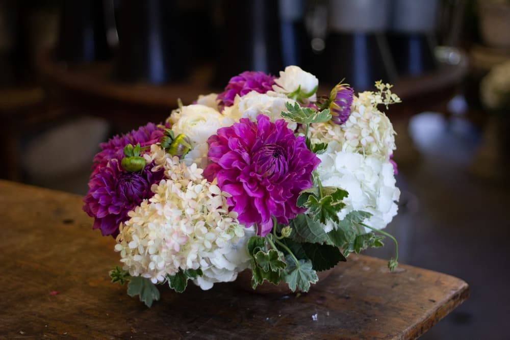 Plum - A combination of full seasonal flowers in purple and white, inside of a round mercury vase. 