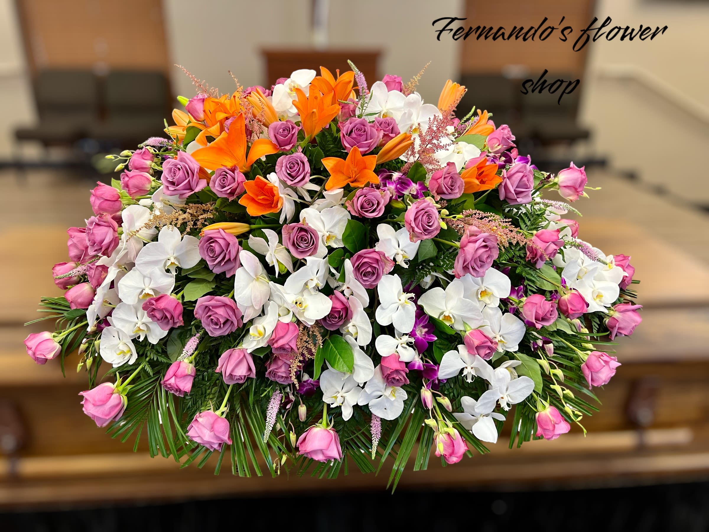 Wings Of Peace By Fernando’s Flower Shop - The image features a stunning floral arrangement atop a wooden casket. The arrangement includes a mix of white orchids, purple and pink roses, bright orange lilies, and lush greenery. The setting appears to be a funeral or memorial service venue, with chairs, a podium, and wooden blinds in the background. The flowers create a vibrant yet elegant tribute.