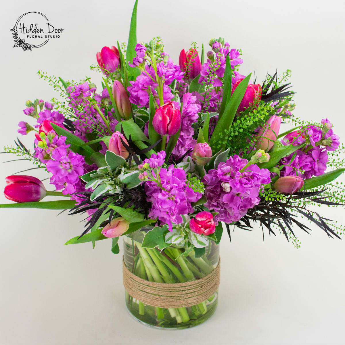 Happiness - Introducing &quot;Happiness,&quot; a radiant floral arrangement that exudes joy and positivity. This delightful bouquet is elegantly presented in a clear glass vase, showcasing the vibrant colors of hot pink stock and hot pink or purple tulips. Please note that the specific shade of the arrangement may vary, adding a touch of uniqueness to each floral creation. The blooms are artfully complemented with seasonal greens, creating a harmonious composition that captures the essence of happiness.  The &quot;Happiness&quot; arrangement serves as a heartfelt reminder of the importance of joy in our lives. This exquisite floral creation is the best choice for various occasions, whether you want to congratulate someone on their graduation, celebrate birthdays and anniversaries, or send get-well wishes to uplift spirits. Its timeless beauty and vibrant hues make it a versatile gift that will make anyone smile.  This classic floral arrangement is artfully designed to enhance any space with its burst of color and positive energy. Place it in a living room, office, or any room that could benefit from a touch of happiness. The vibrant hot pink stock and tulips, accompanied by seasonal greens, create a visually stunning floral display that brightens the surroundings and creates a sense of joy.   