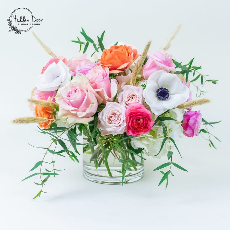 Vivaldi - I Prepare to be captivated by the true masterpiece of color and cheerfulness that is the &quot;Vivaldi&quot; floral arrangement. This contemporary centerpiece is a delightful ensemble of anemones, ranunculus, and a vibrant mix of large and spray roses, all coming together in a stunning garden of flowers. Each &quot;Vivaldi&quot; arrangement is a unique work of art, showcasing slight color and shade variations that make it a one-of-a-kind beauty. No two arrangements are alike, ensuring the recipient receives an exceptional and individual creation. While they may differ, one thing is sure: they will all exude an exquisite beauty that will leave a lasting impression. Radiating happiness, cheerfulness, and whimsy, the &quot;Vivaldi&quot; floral arrangement is a perfect present for anyone in your life. Whether it's a cherished family member, a dear friend, a romantic interest, or a valued business partner, this arrangement will bring a touch of joy and add a harmonious note to their day. Just as a symphony evokes emotions and creates beautiful melodies, the &quot;Vivaldi&quot; arrangement aims to bring a moment of happiness and brighten the recipient's life. Its vibrant colors, and playful composition will transport them to a whimsical garden, where every petal dances to its own rhythm.  