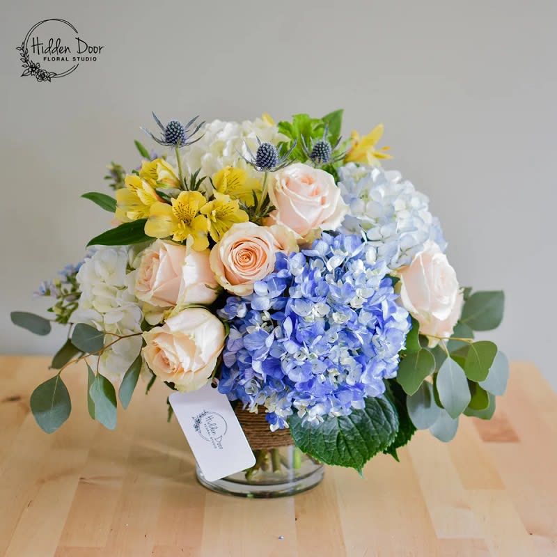 Catalina -   The &quot;Catalina&quot; arrangement features the radiant yellow of alstroemeria, symbolizing the warmth and brilliance of the sun. These vibrant blooms bring a feeling of joy and energy, infusing the arrangement with a radiant glow that uplifts the spirits and adds a touch of sunshine to any space. Complementing the alstroemeria are the serene blue hydrangeas, reminiscent of the endless expanse of the ocean. These blooms evoke a sense of tranquility and calm, transporting the viewer to the soothing embrace of coastal waters. Their delicate petals mirror the ebb and flow of the tides, creating a mesmerizing visual experience. The arrangement incorporates roses in a rich brownish hue to represent the sandy shores. These blooms bring a touch of warmth and earthiness to the design, mirroring the soft grains of sand that define the island's beaches. They add depth and contrast, harmonizing the colors of the sun and the sea.  The &quot;Catalina&quot; arrangement is presented in a transparent, clear vase elegantly adorned with jute wrapped around it. This design choice not only complements the maritime theme but also allows the vibrant colors of the flowers to take center stage, creating a captivating visual impact.    