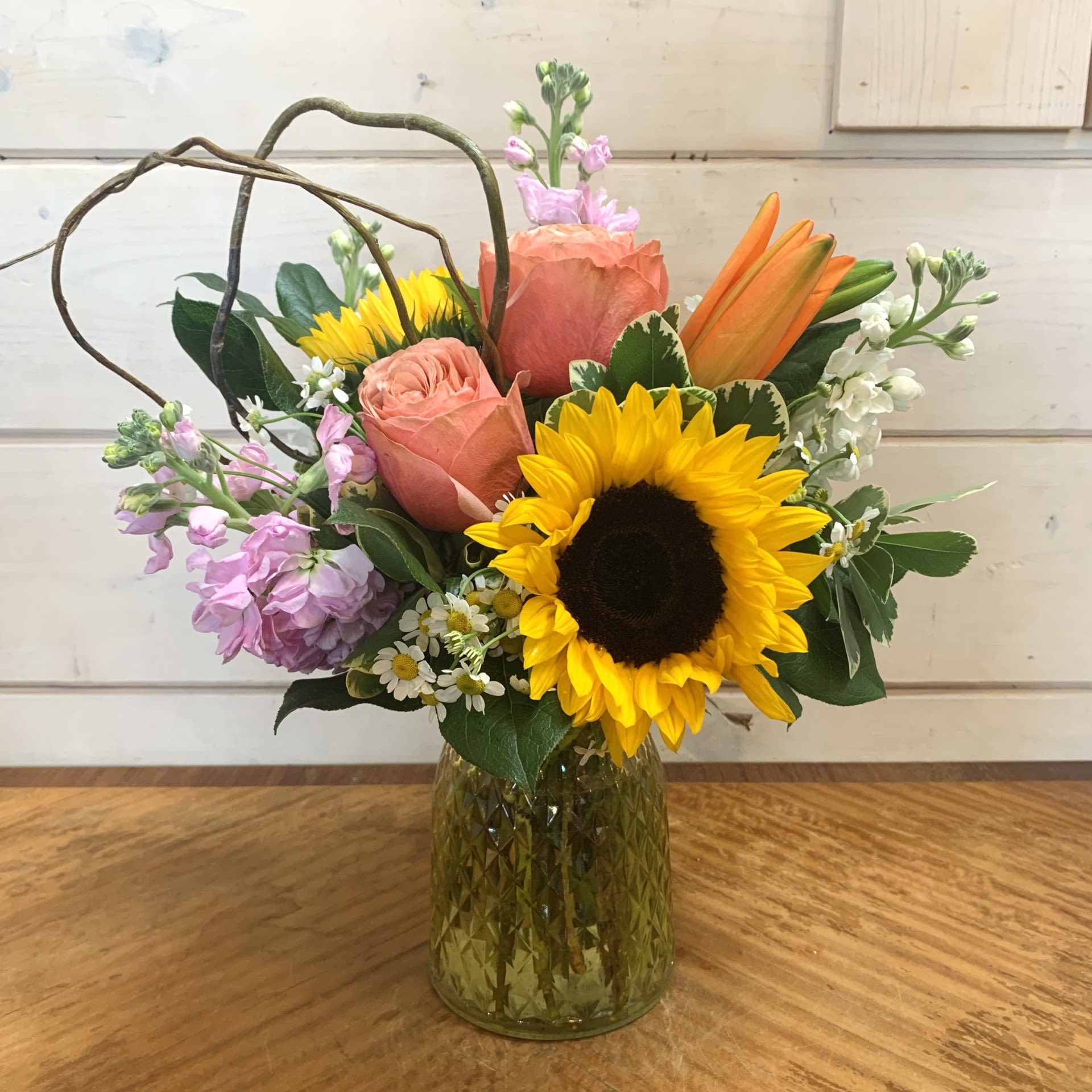 Pollinator's Paradise - Sunflowers and apricot roses with an orange lily, and fragrant pink and white stock arranged in a yellow tinged cut glass posy vase with chamomile and curly willow. Approximately 14&quot; tall and 12&quot; wide.