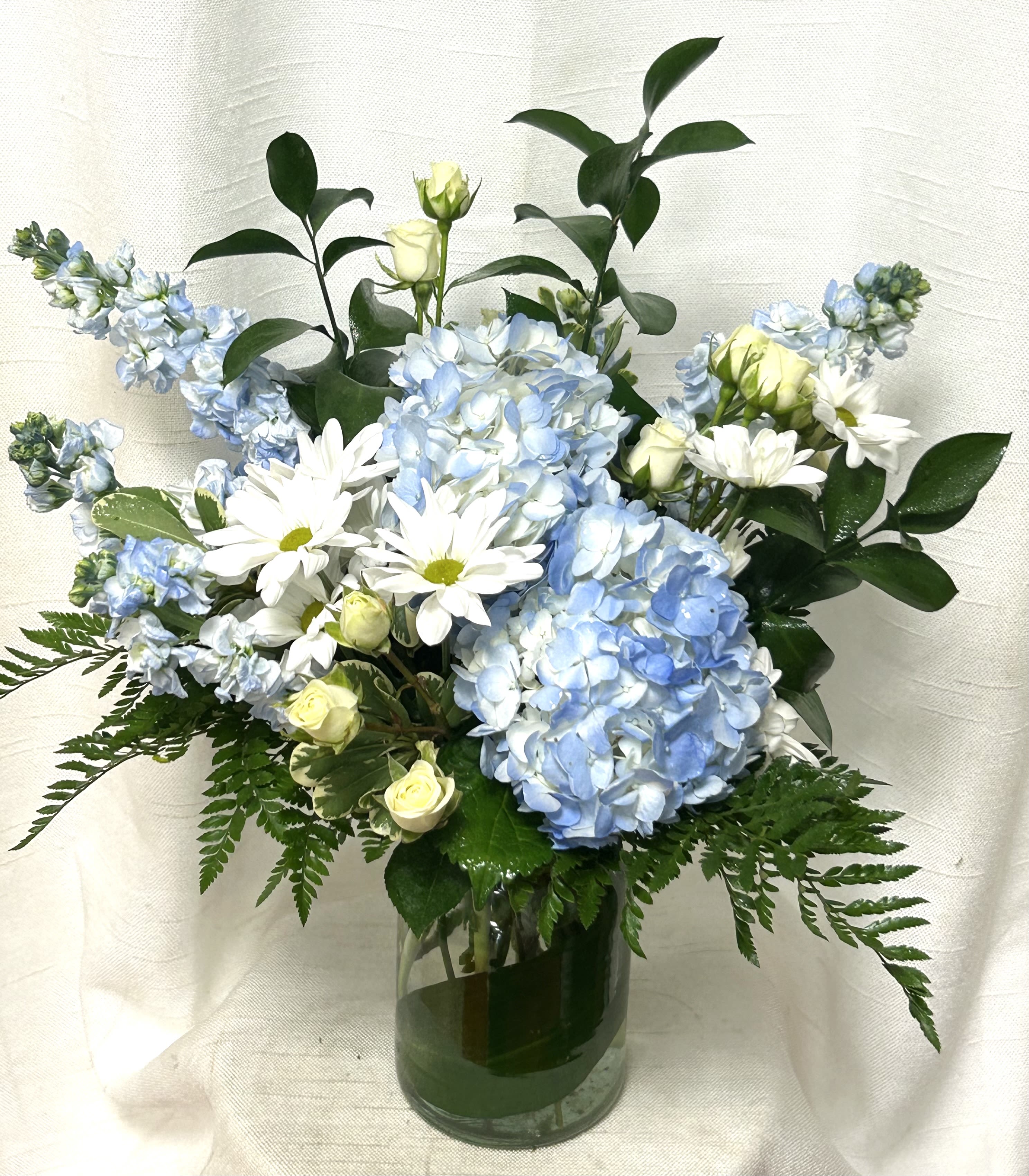 Sky Blue (BB) - Immerse yourself in a tranquil oasis with our enchanting clear skies flower arrangement, showcasing a harmonious blend of blue hydrangeas, white roses, blue delphiniums, and white poms. Artfully arranged in a leaf-lined glass cylinder vase, this stunning ensemble evokes a sense of serenity and beauty.