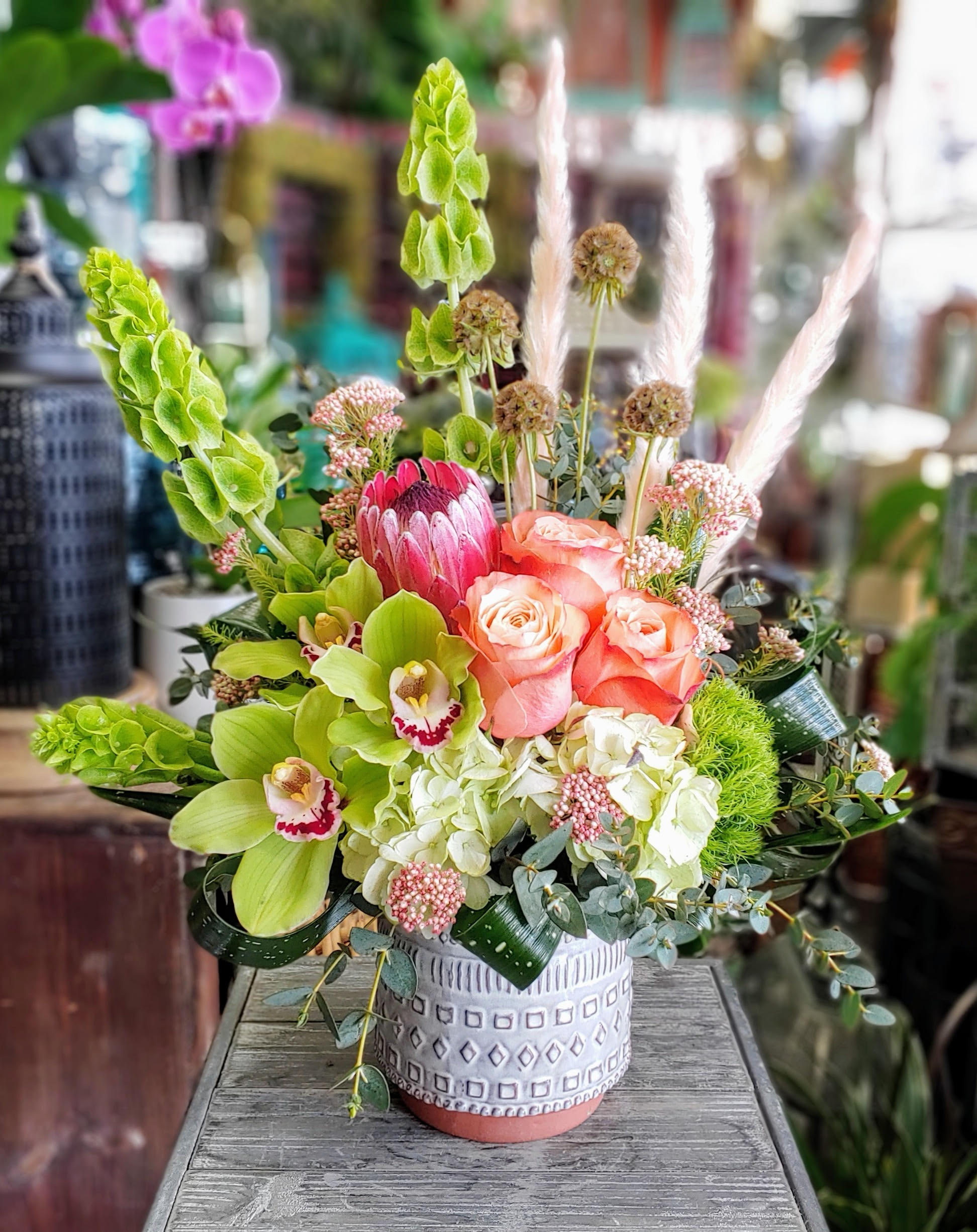 Gypsy Gypsy - Gypsy Wildly elegant and full of untamed charm, Gypsy is a striking floral arrangement from Windmill Floral Expressions in Las Vegas, designed for the bold and the bohemian at heart. Bursting with vibrant hues and eclectic textures, this piece feels like a floral journey—free-spirited, earthy, and full of magic. At the center, a stunning pink protea stands proud, encircled by peach roses, lime green cymbidium orchids, and soft green hydrangeas. Wisps of blush pampas grass and tall bells of Ireland add height and movement, while scabiosa pods, green trick, rice flower, and glossy tropical foliage create layers of visual intrigue. Presented in a modern ceramic pot with geometric patterns, Gypsy makes a captivating gift or showpiece—perfect for those who live colorfully and love deeply. Handcrafted with love in Las Vegas, NV. Same-day delivery available.