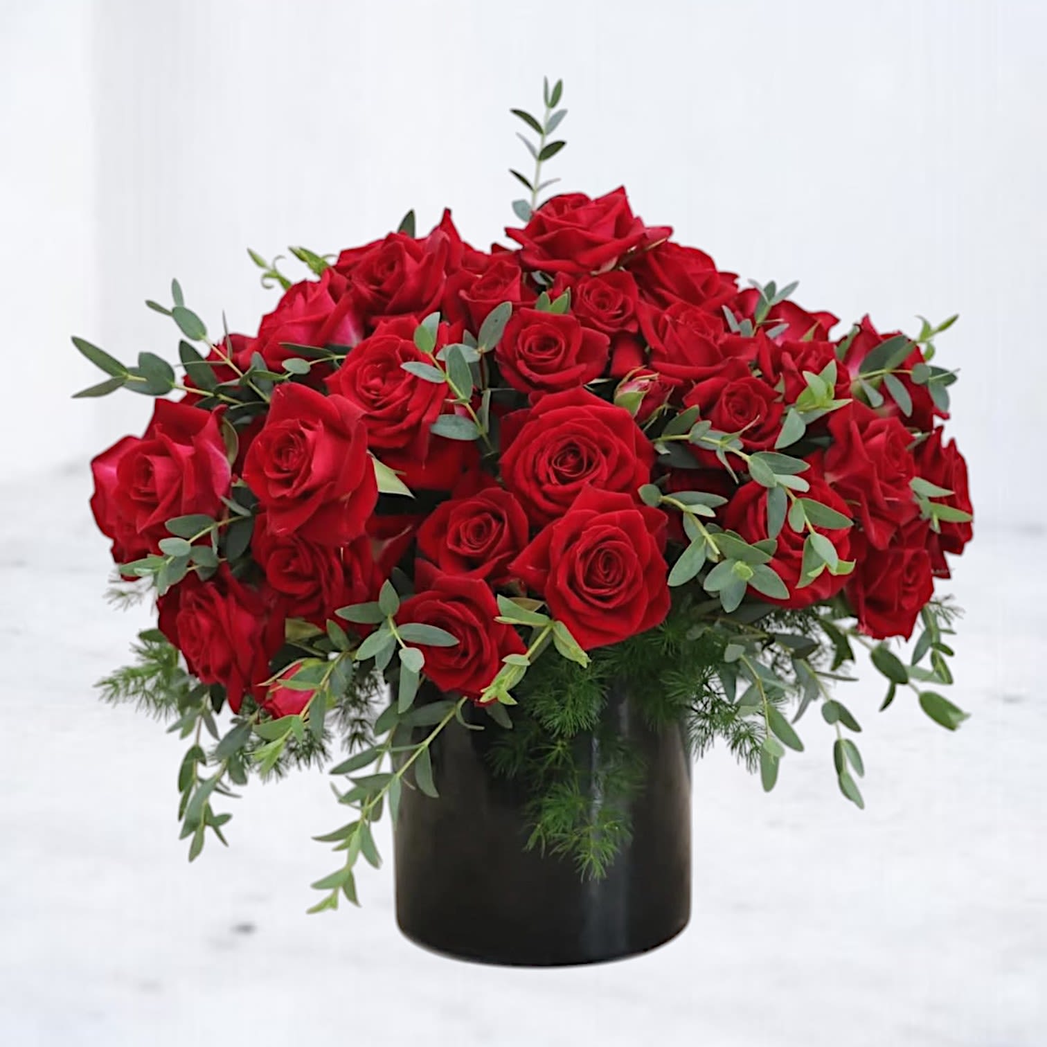 The Iconic Red - Make an unforgettable statement with this bold and breathtaking arrangement of premium long-stem red roses, artfully designed in a sleek black vase. Overflowing with 48 velvety red roses and lush greenery, this luxurious bouquet radiates passion and sophistication.  Perfect for grand romantic gestures, milestone anniversaries, or when only the most stunning roses will do, this modern design brings a dramatic edge to a timeless classic. The black vase provides striking contrast to the vivid blooms, creating an arrangement that turns heads in any setting.  Send this show-stopping rose arrangement for same-day delivery in Houston, Galveston, and surrounding areas — an unforgettable gift of love, admiration, or celebration.  Features:  48 premium long-stem red roses Designed with lush seasonal greenery (may include eucalyptus and fern) Arranged in a black cylinder vase Available for same-day delivery in Houston, Galveston, The Woodlands, Magnolia, Conroe, Spring, and nearby communities Substitution Policy: In rare cases, exact greenery varieties may vary due to seasonal availability, while maintaining the overall design, style, and premium quality of the arrangement. Vase style and color will remain consistent as shown.
