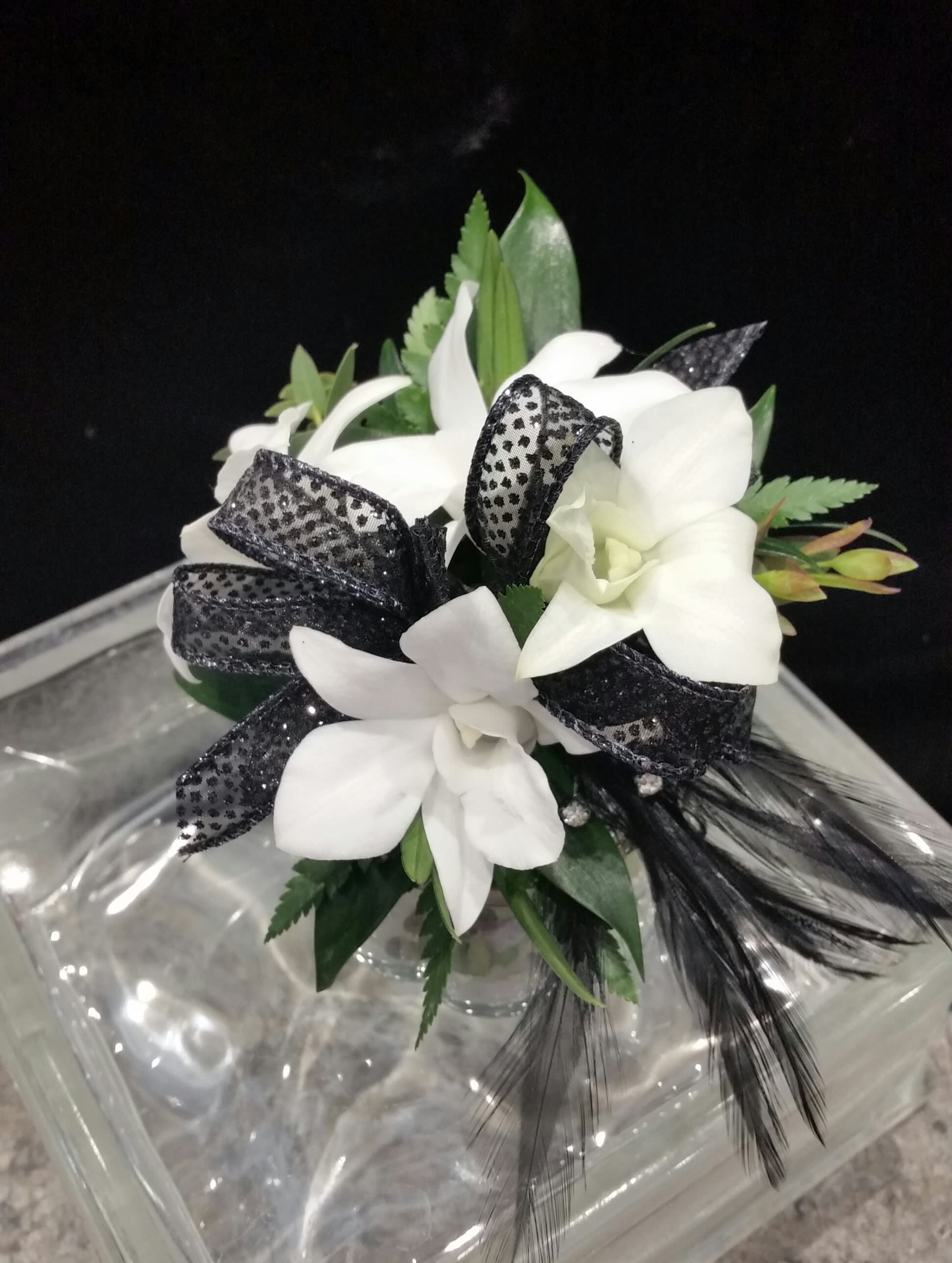 'Dottie' black and white wrist corsage 'Dottie' black and white wrist corsage - Perfect, delicate, white dendrobium orchids amongst a bed of greens and a simple dotted swiss black ribbon. Simple, beautiful, elegant. And for a daring design accent, we've added rhinestone studded black feathers.