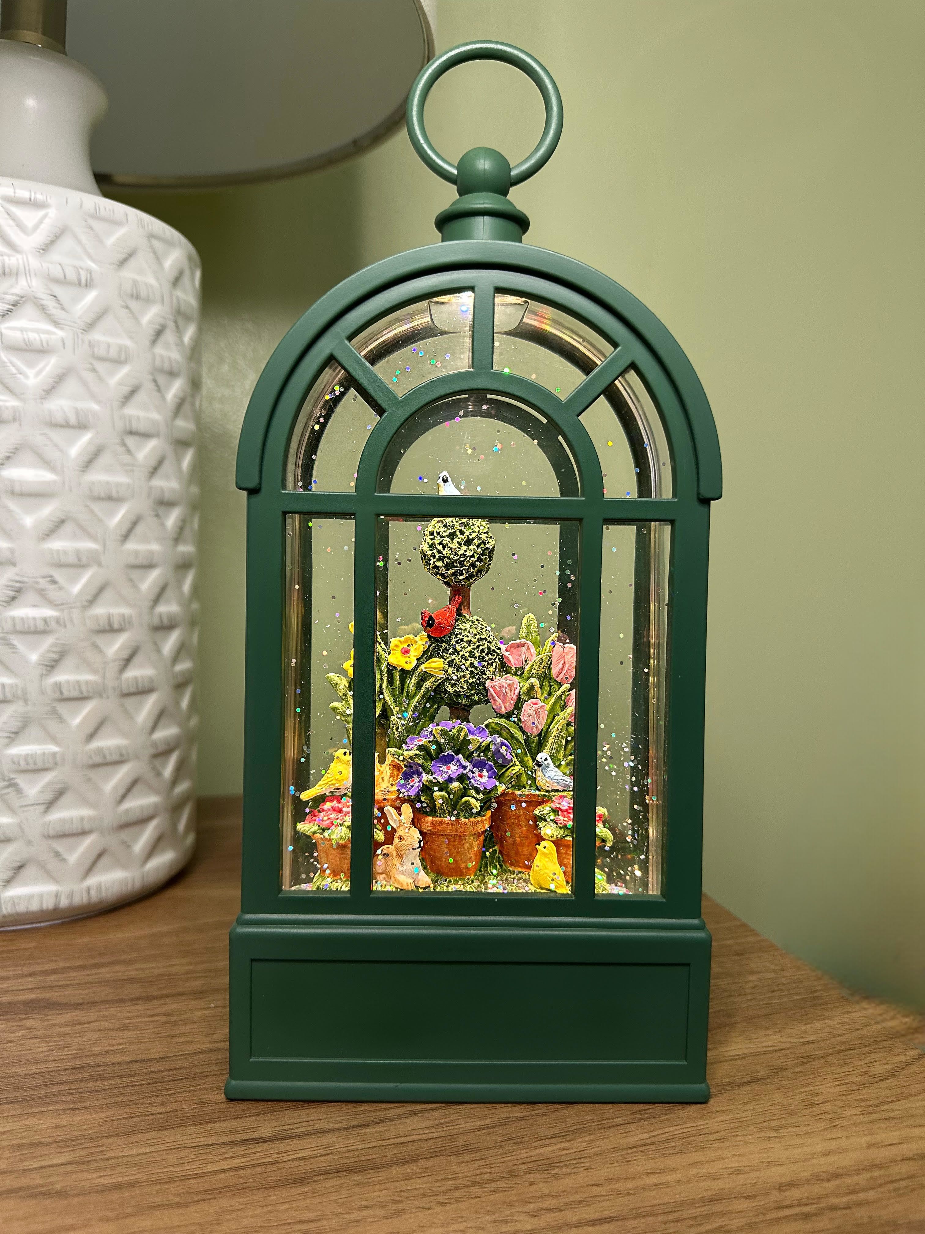 Spring-themed Snow Globe Lantern. Spring-themed Snow Globe Lantern. - "I am a charming, green metal lantern with a glass enclosure, housing a whimsical spring scene. Inside, a miniature garden is teeming with life, featuring potted flowers, birds, and bunnies. The lantern is adorned with glittering particles that resemble snow or sparkles, adding to its enchanting ambiance. I sit atop a wooden table, surrounded by a soothing green wall and a white decorative object, exuding a sense of serenity and joy.