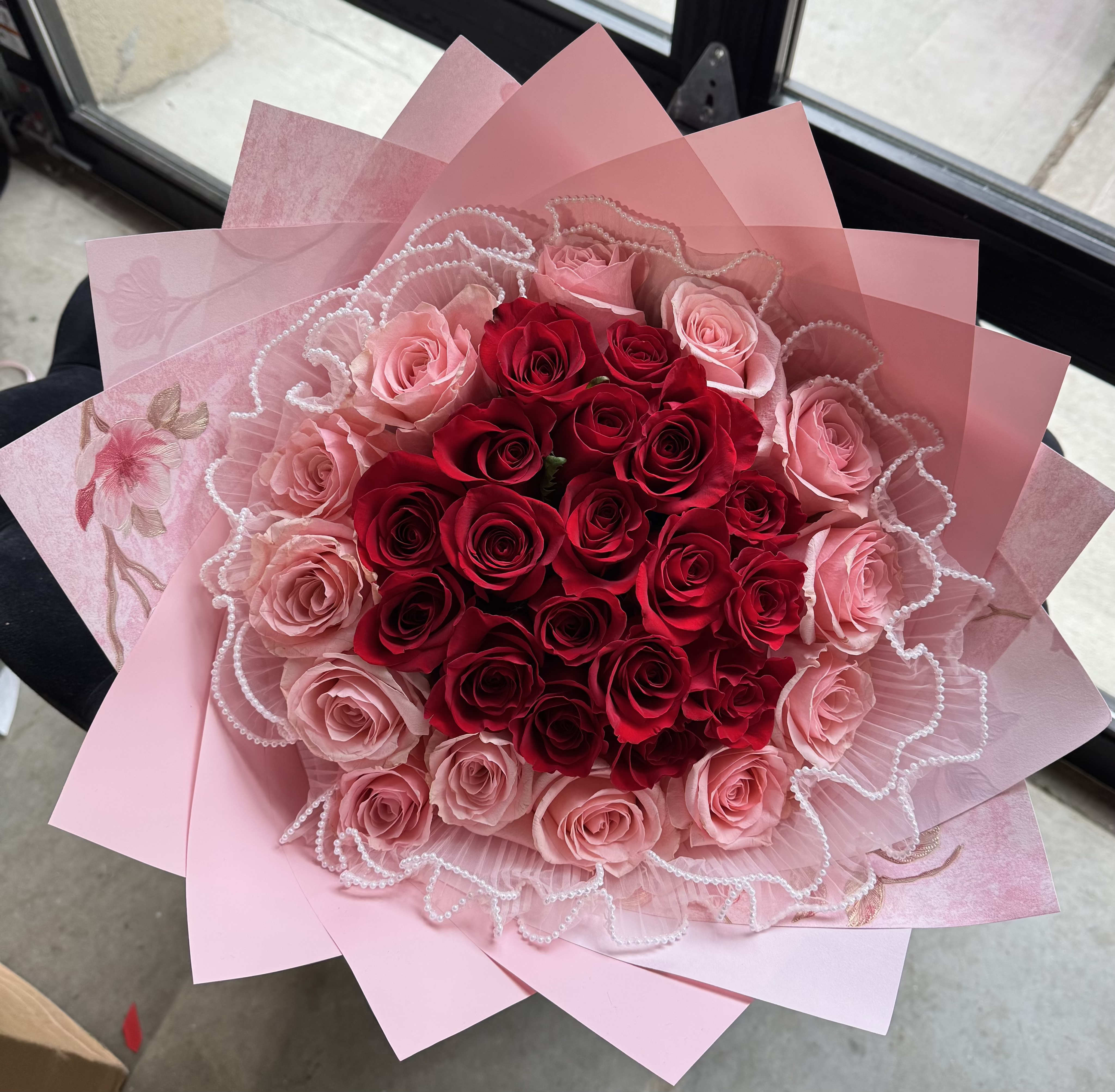 Blushing Heart Rose Bouquet - A classic expression of deep affection and timeless grace, the Blushing Heart Rose Bouquet features a lush cluster of passionate red roses at the center, beautifully surrounded by delicate blush pink roses. Nestled in layers of soft pink wrapping and trimmed with pearl garland, this bouquet radiates romance and refinement — perfect for anniversaries, proposals, or any heartfelt occasion.