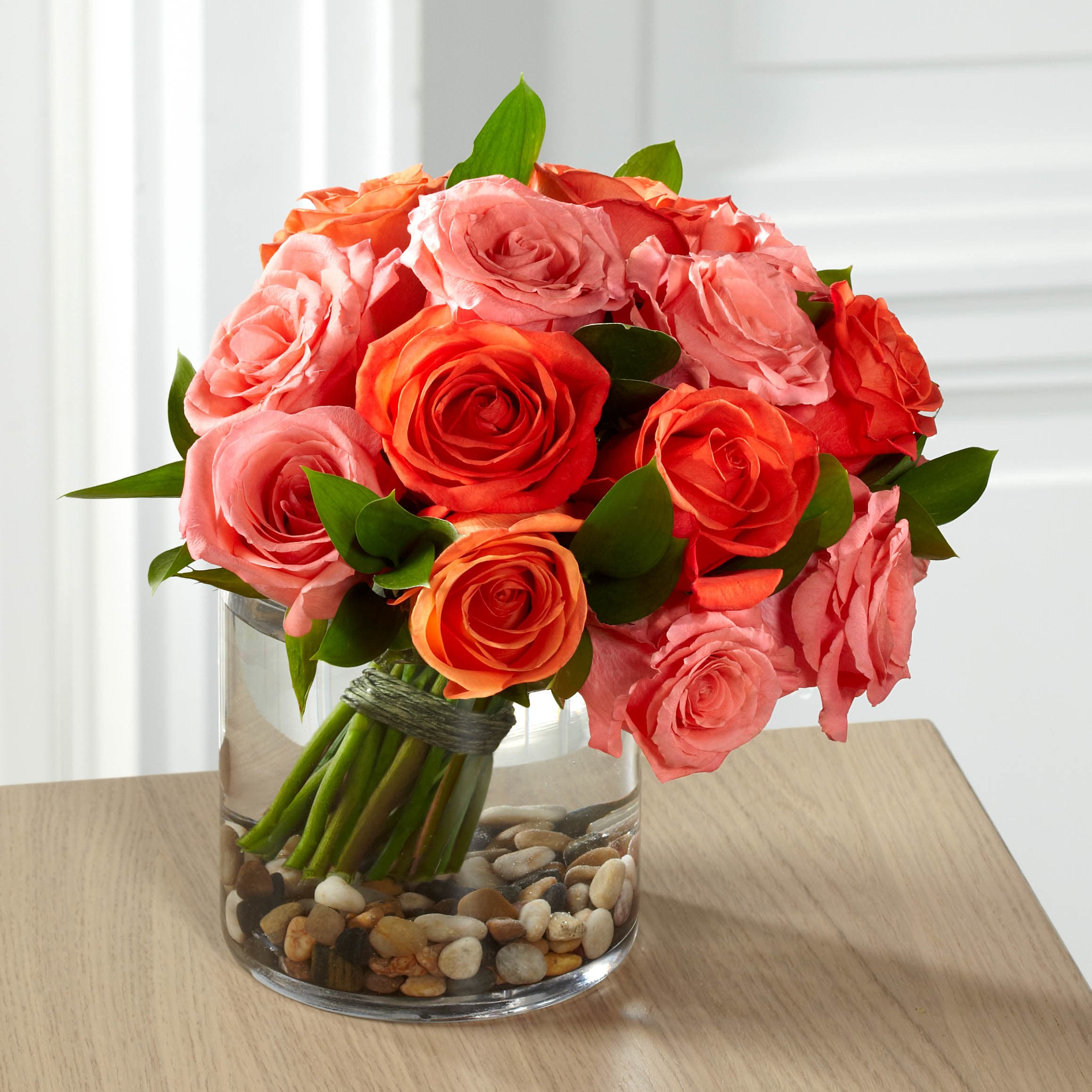 E8-5235 - Pebble Beach Bouquet - Blooming with grace and style, this stunning rose bouquet is set to send warm wishes to your recipient in way that will lift their spirits and have them delight in the unexpected. Salmon roses and coral roses combine to create a bright blushing display, accented with lush greens and hand-tied to provide a rounded and full form. Casually placed in a large clear glass cylinder vase with river rocks at the bottom to create even further interest.
