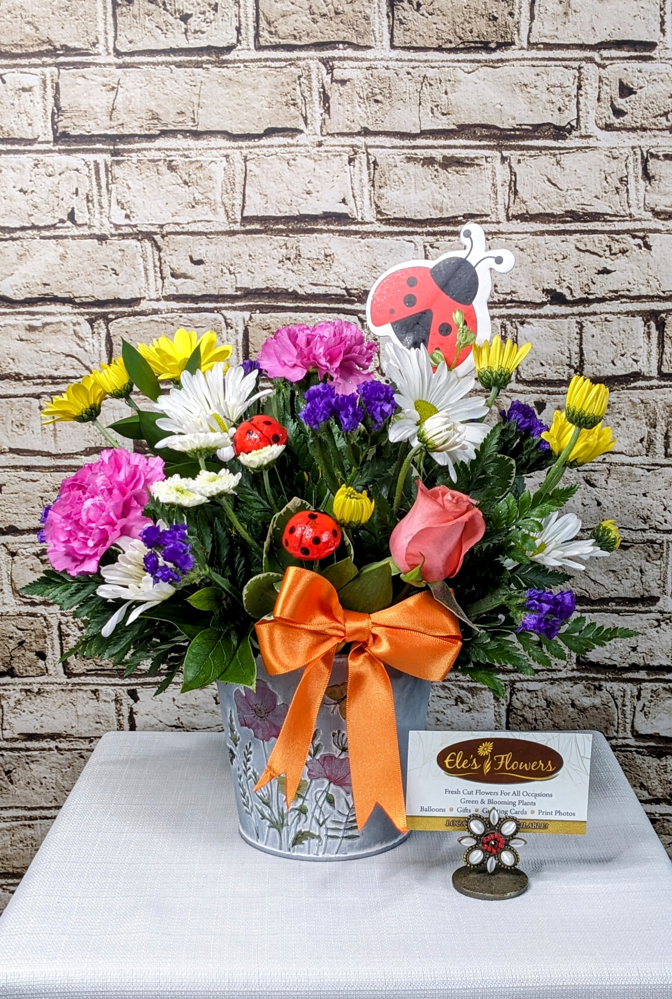 Ladybug - Bursting with garden-fresh charm, Ladybug is a whimsical arrangement that’s as fun as it is bright! A lively mix of pink carnations, white daisies, sunny yellow mums, purple statice, and a single sweet rose is tucked into a wildflower-inspired metal pot, finished with a cheerful orange bow. Topped with playful ladybug accents—including a big smiling one ready to fly—this design is sure to bring smiles, sunshine, and a touch of luck to any day.  Perfect for birthdays, just-because days, or anyone who could use a little joy and whimsy!  Approximate Dimensions:  14&quot;H x 12&quot;W