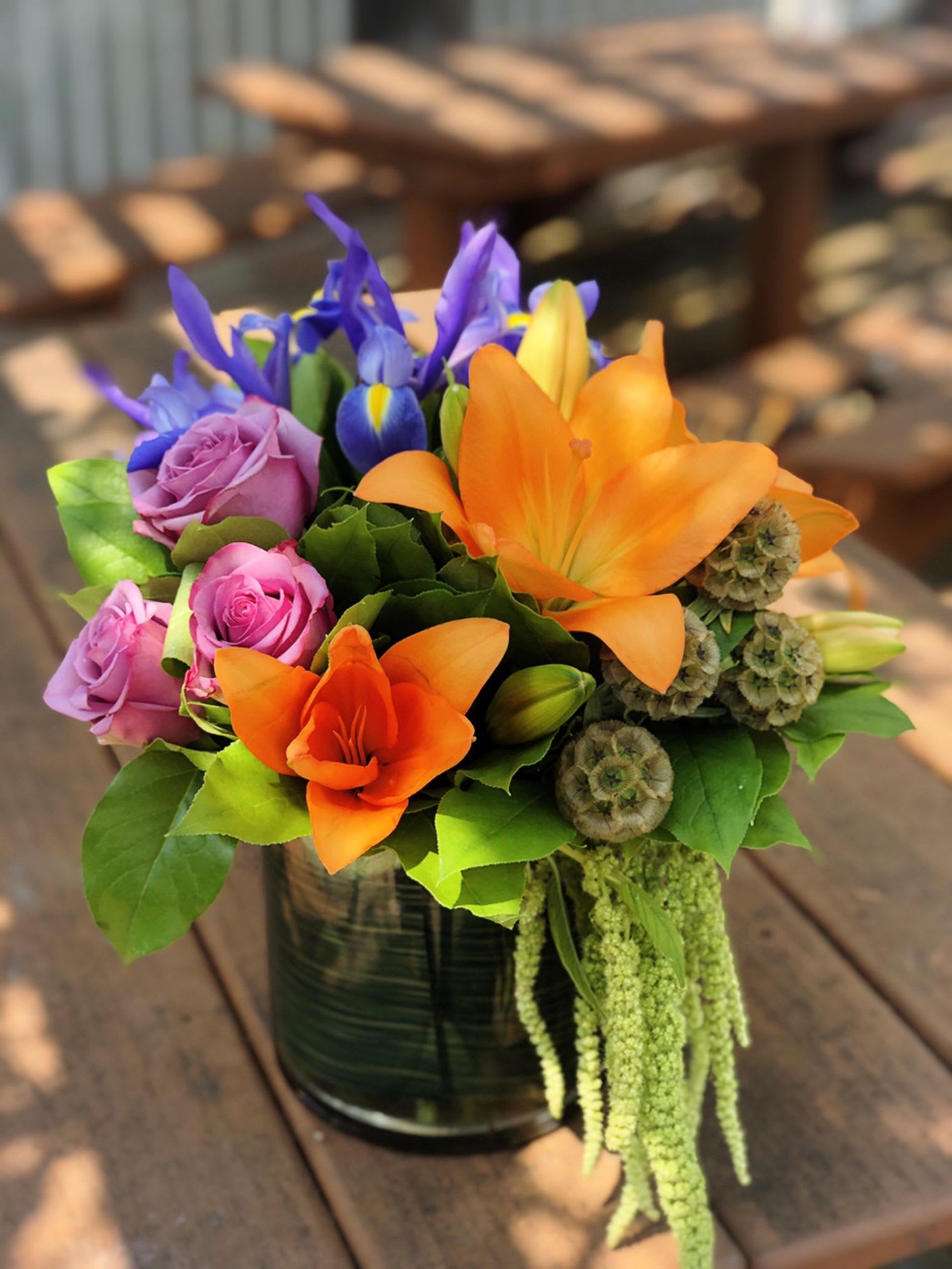 Colour Burst Colour Burst - -3 Stems Orange Lilies -6 Stems Purple Roses -3 Stems Blue Iris - Amaranthus -Greenery -6x6 Cylinder Vase