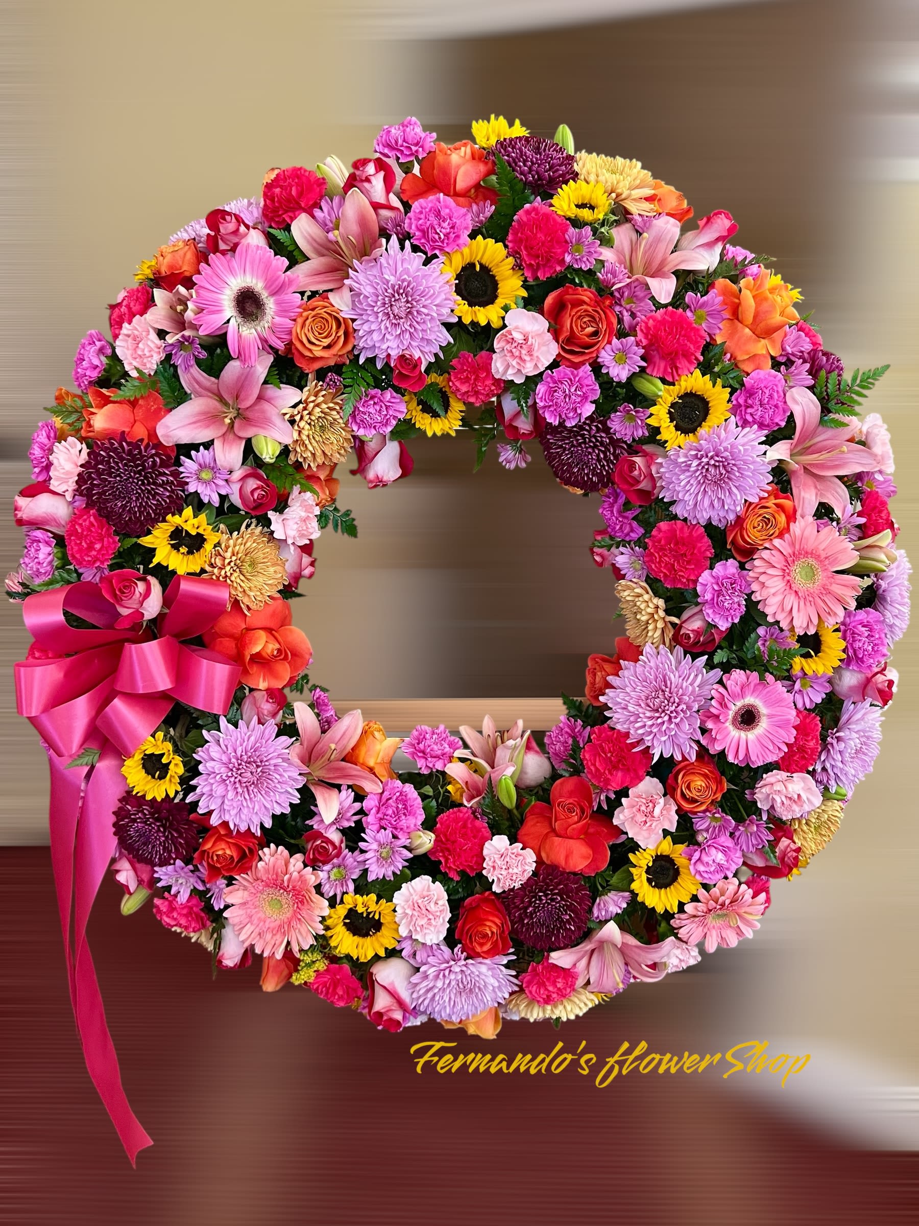 Until The End - This is a vibrant and colorful floral wreath designed to celebrate life and joy. It features an eclectic mix of flowers in shades of pink, red, orange, yellow, and purple. The arrangement includes sunflowers, lilies, roses, gerbera daisies, and chrysanthemums, symbolizing warmth, happiness, and love. The addition of lush greenery enhances the bold colors, while a striking pink ribbon adds an elegant finishing touch. Perfect for cheerful events or heartfelt tributes, this wreath radiates beauty and positivity.