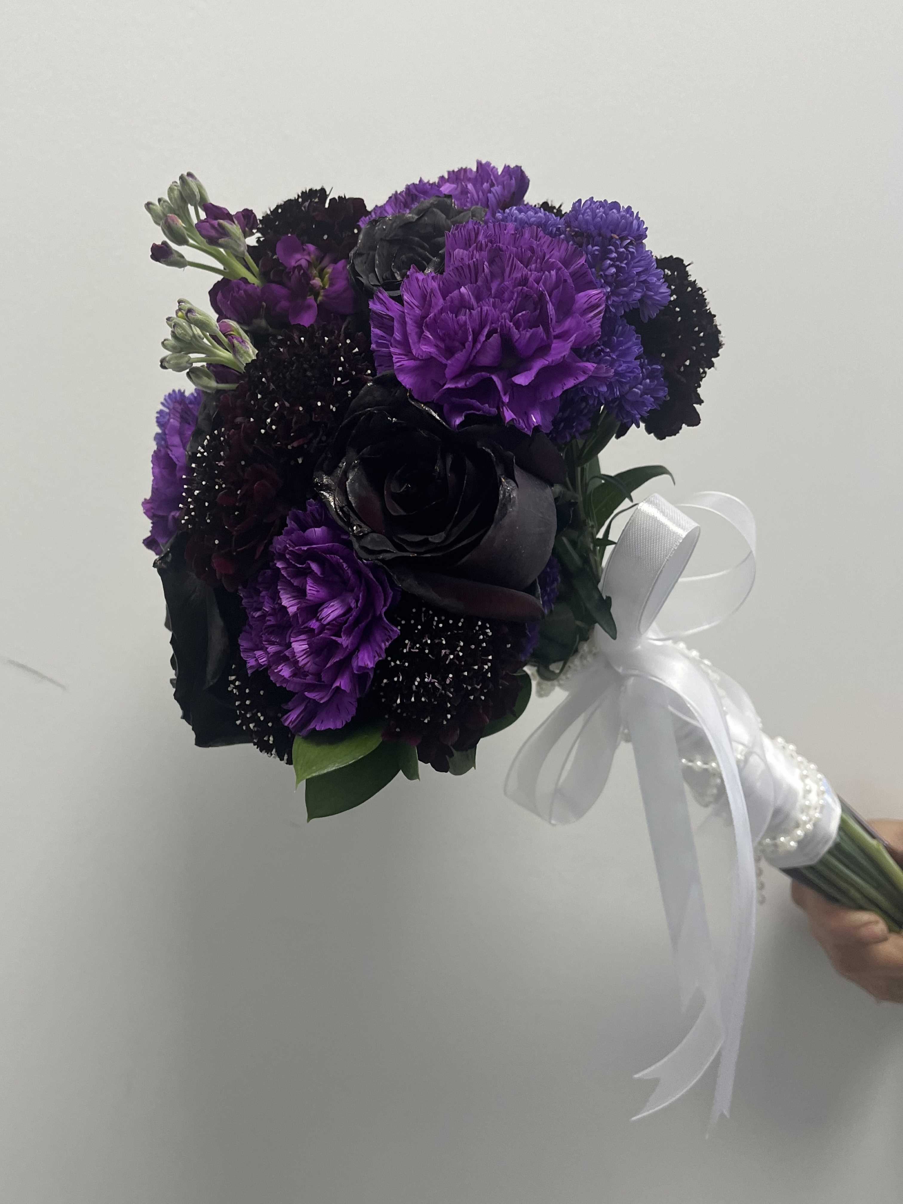 Twilight Purple Twilight Purple - Features purple carnations, black roses, lavender stock, and purple scabiosa wrapped in a white ribbon with pearls.