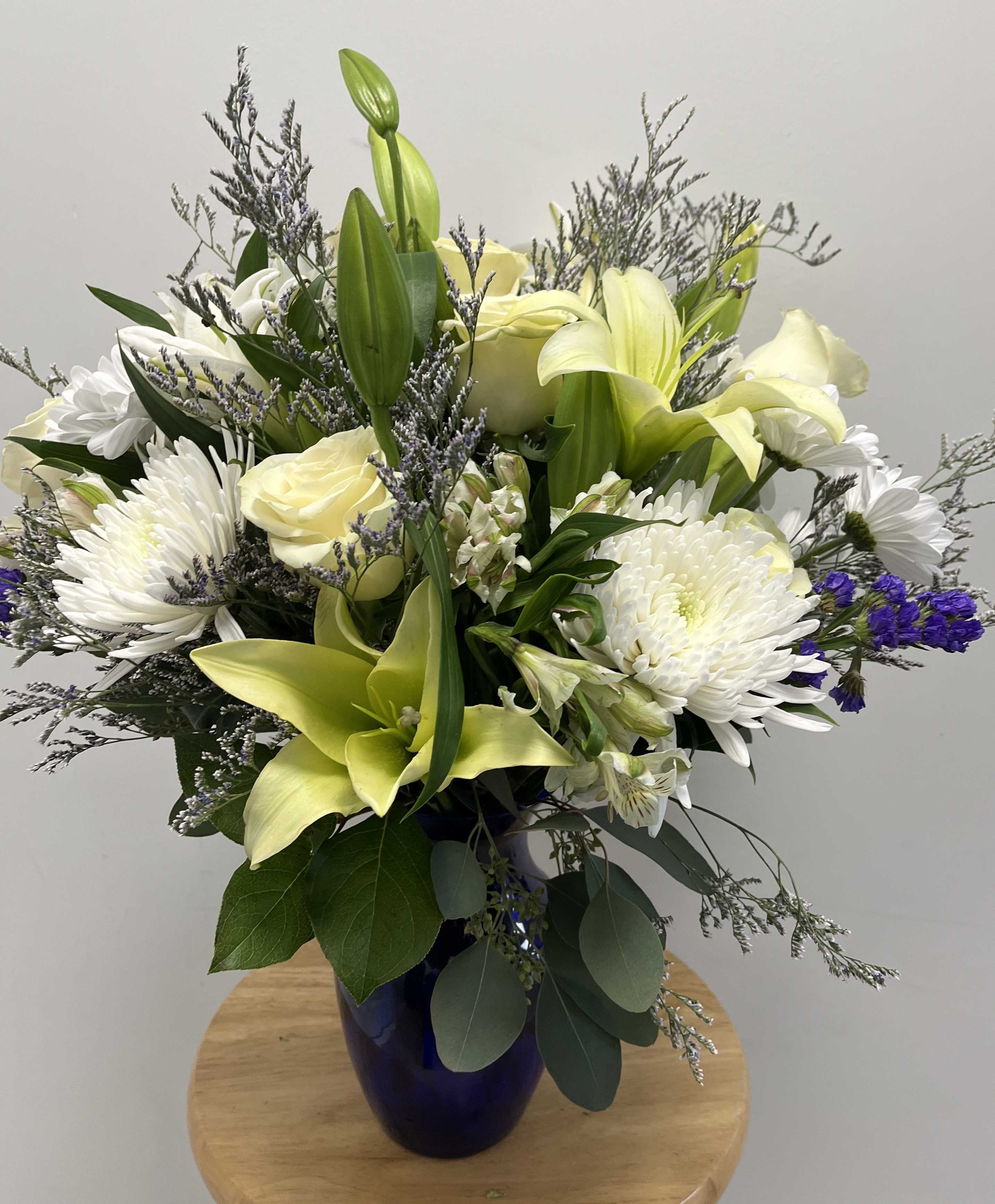 Elegant Winter Bloom - This arrangement features a blend of white roses, white lilies, white chrysanthemums, sprigs of purple statice, accented by silver-toned filler and greenery. Displayed in a deep blue vase, it is perfect for formal and heartfelt occasions. 