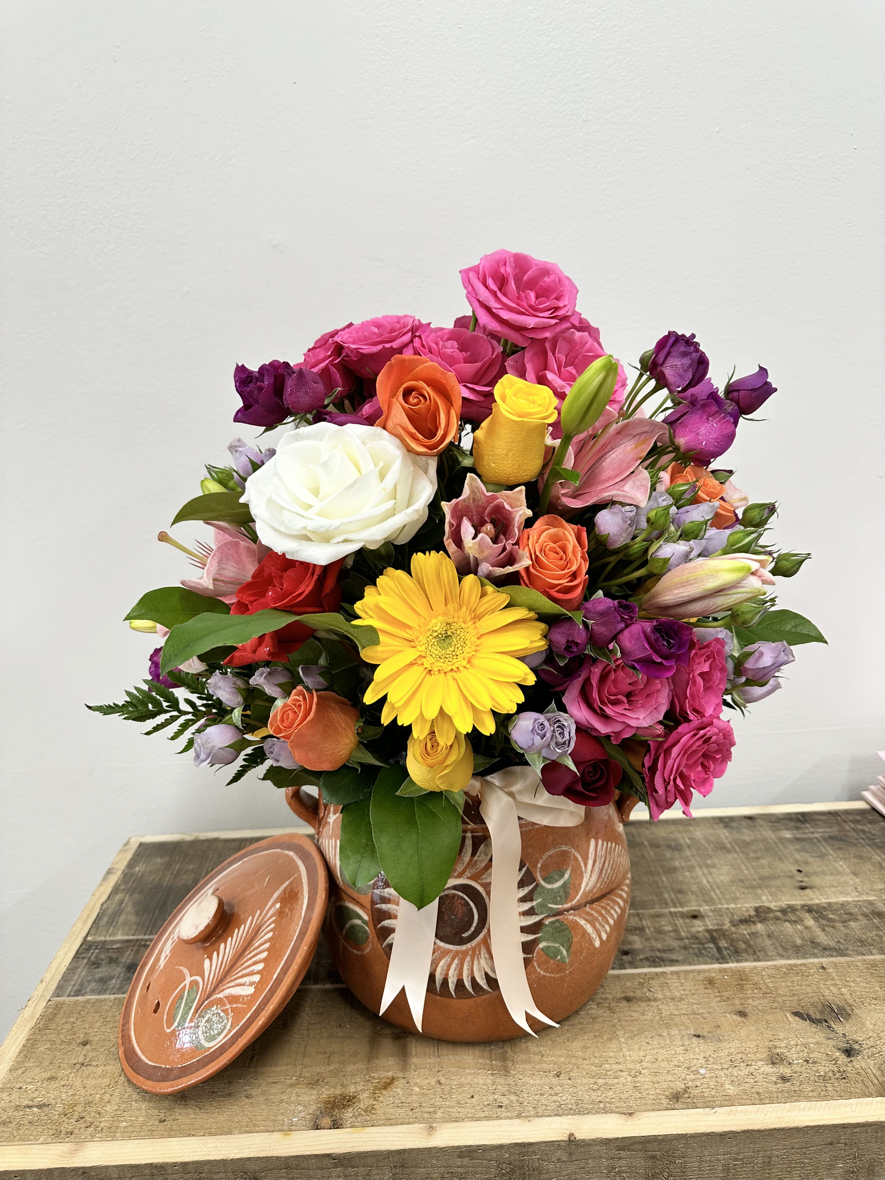 Artisans Bloomd -  Nestled in a handcrafted clay pot, this vibrant bouquet features an enchanting mix of pink, white, red, and orange roses, complemented by delicate light purple and lavender spray roses.