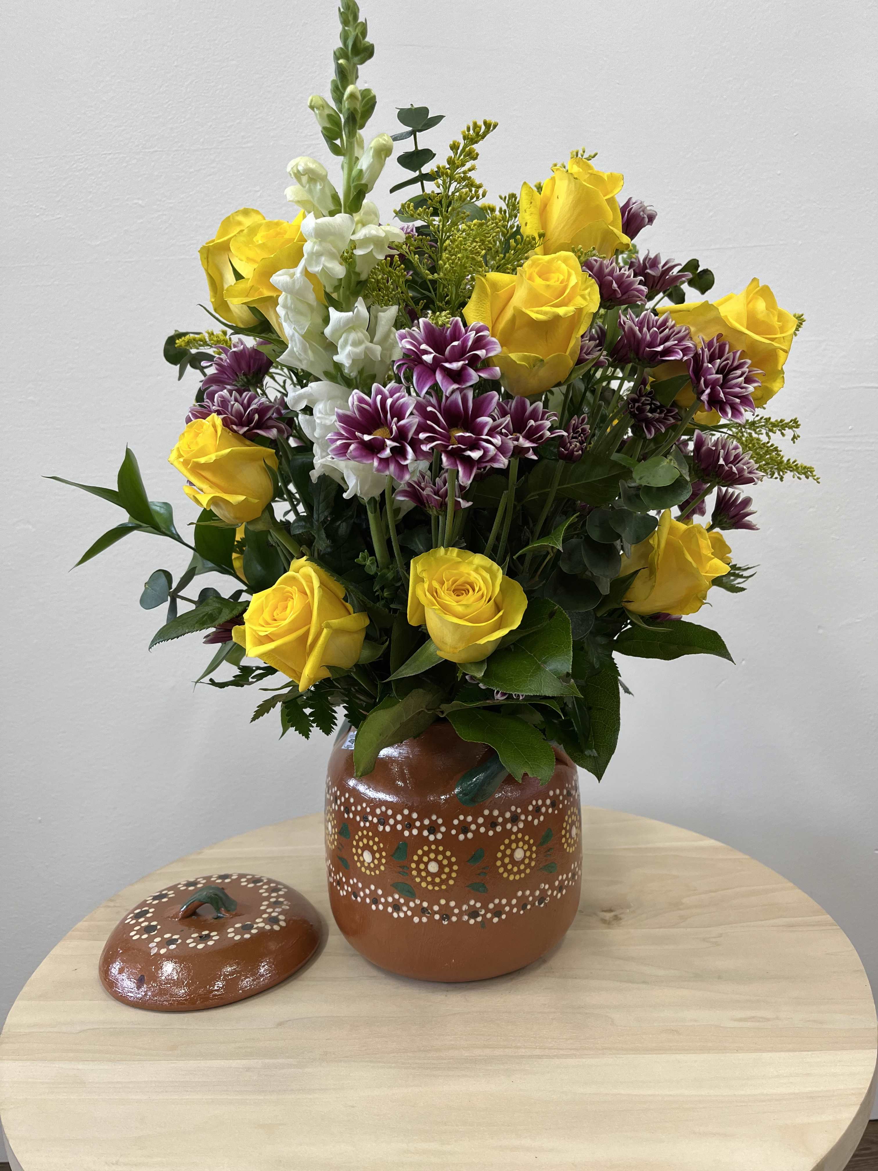 Dulce De Encanto - A cheerful mix of yellow roses, white stock, and purple daisies in a rustic clay pot. Perfect for adding a touch of color and charm.