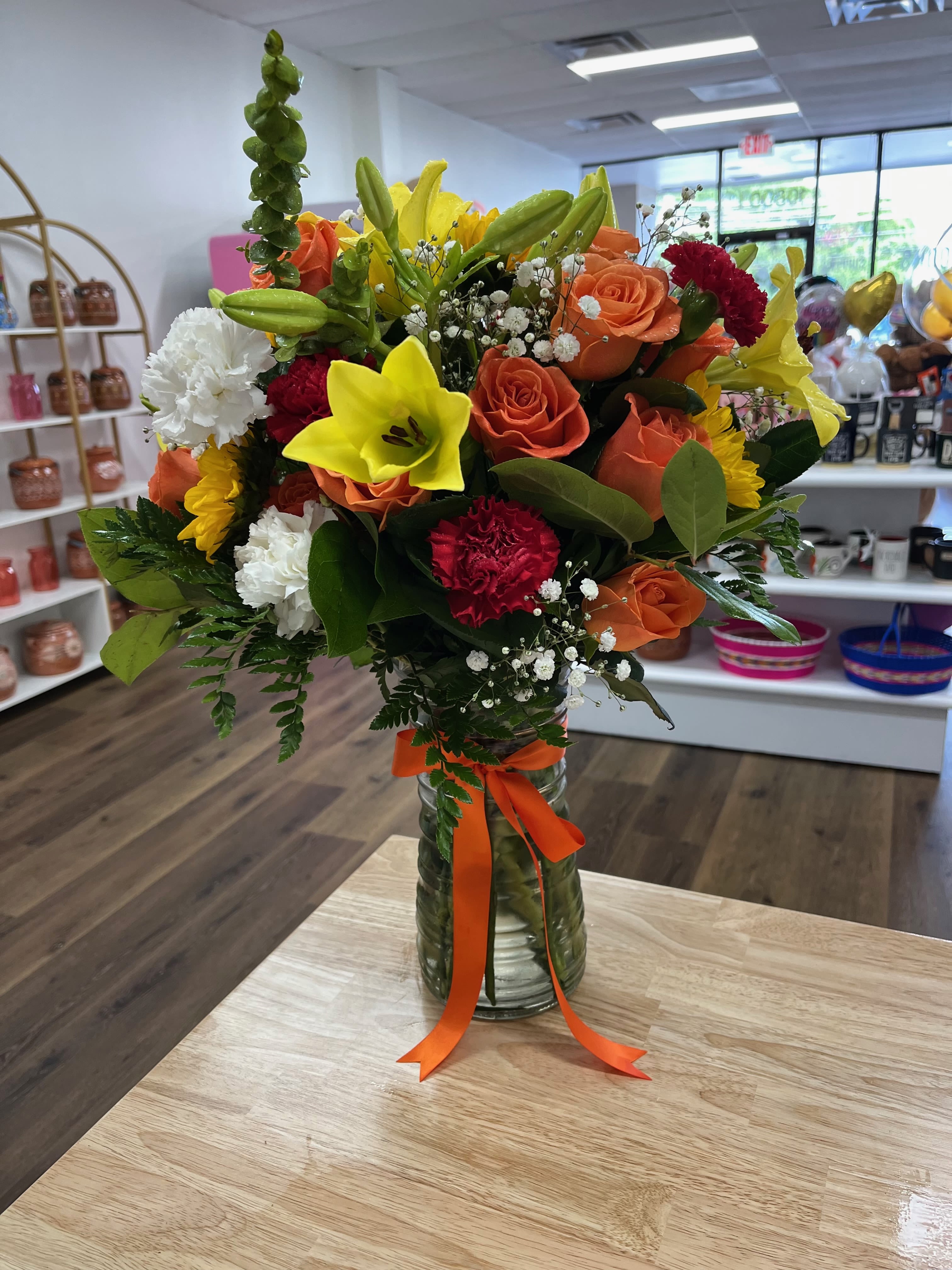Vivid Symphony  - Orange roses, red, white, and yellow carnations, and bright yellow lilies are beautifully complemented by green stocks and lush greenery. Arranged in a tall glass vase, this striking bouquet brings a lively burst of joy and warmth to any setting.