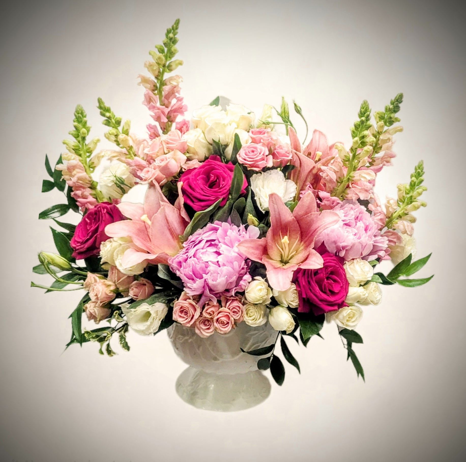 blushing Elegance blushing Elegance - Delight in the soft romance of our Blushing Elegance, a luxurious arrangement overflowing with pink lilies, vibrant roses, lush peonies, delicate spray roses, snapdragons, and lisianthus. Artfully arranged in a classic white pedestal vase, this bouquet exudes charm and grace, making it a stunning gift for weddings, anniversaries, Mother's Day, or simply to express love and admiration. Approximate Size: 18" W x 16" H Flower Types: Pink lilies, roses, peonies, spray roses, snapdragons, lisianthus, assorted greenery Presentation: Arranged in an elegant white footed vase Ideal For: Romantic occasions, Mother’s Day, Weddings, Birthdays, Just Because