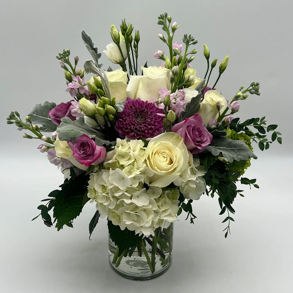 PICTURESQUE - Gorgeous flower arrangement adorned with unique and different flowers such as white and purple roses, white Hydrangeas and delicate white Lisianthus, tall white and lavender stock flower with a touch of silvery dusty miller and fragrant eucalyptus parvifolia, all in a crystal clear  vase.   