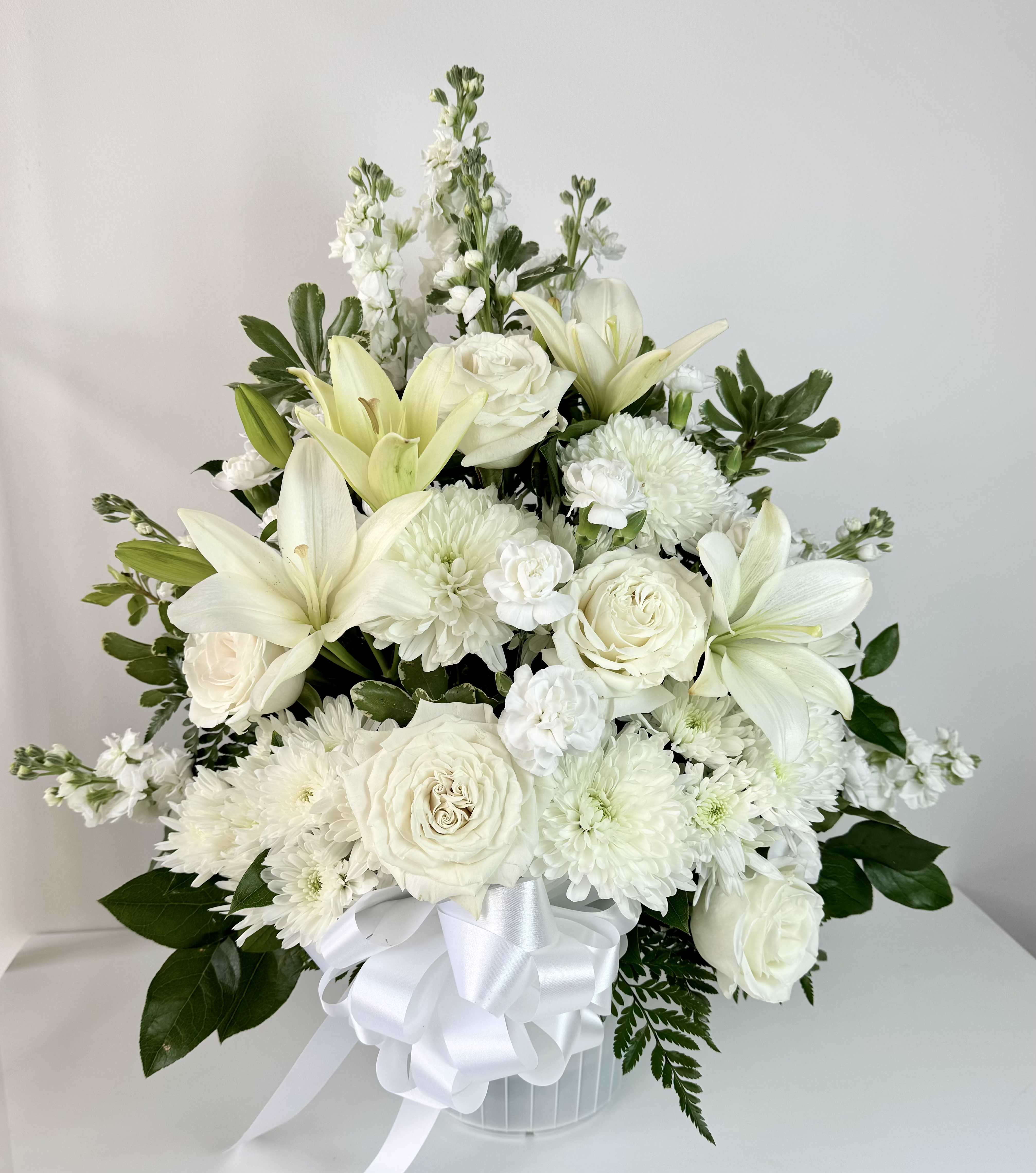 Heavenly Grace Sympathy Arrangement - Express your heartfelt condolences with this elegant sympathy arrangement, designed to bring comfort and peace. Featuring serene white lilies, classic roses, delicate spray roses, graceful alstroemerias, and soft pompon carnations, this bouquet offers a gentle reminder of love and remembrance. Beautifully arranged with fresh greenery, it’s a meaningful tribute for a memorial service, funeral, or to send directly to grieving loved ones.  Available in all-white or soft pastel tones. Custom ribbon messages available at Checkout