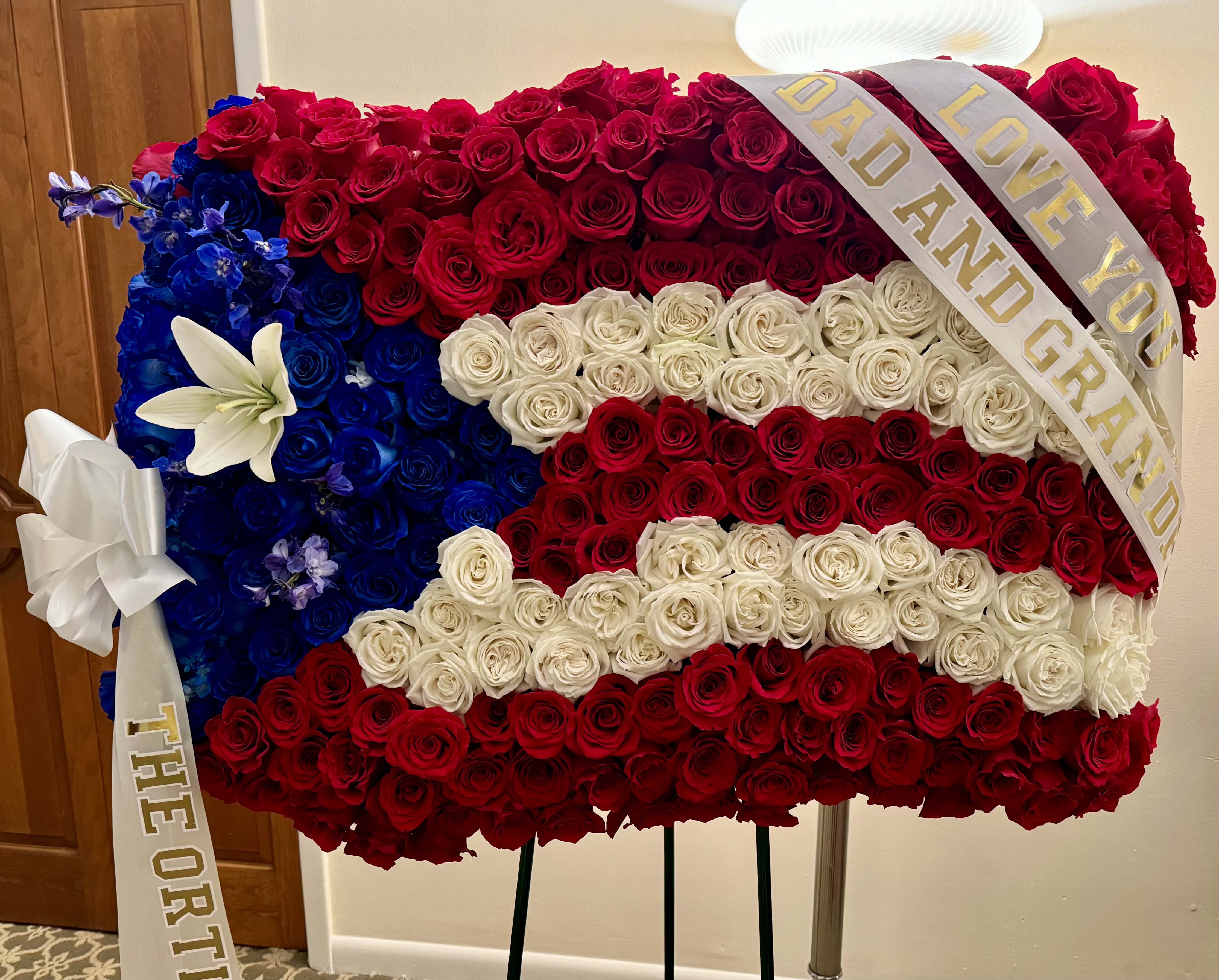 Patriotic Tribute Standing Spray - Honor a life of service and strength with this flag-inspired standing spray. Designed in the colors of the Puerto Rican flag, it features rich red, white and blue roses and vibrant blue delphiniums. This striking arrangement is a meaningful tribute to anyone who proudly represented their country. Displayed on a standing easel, it’s a bold and heartfelt expression of gratitude and remembrance.  Custom ribbons and service emblems available upon request. 