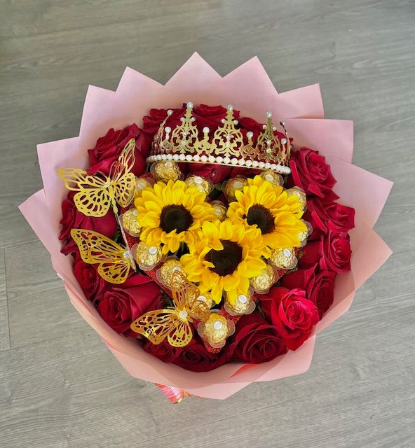 Sweet Majesty - A luxurious heart-shaped arrangement, sculpted with care and overflowing with meaning. The outline of the heart is formed with a lush bed of rich red roses, their velvety petals curling open in perfect harmony, symbolizing deep love and affection.  At the center of the arrangement sits a chocolate heart—smooth, glossy, and nestled like a sweet treasure. It’s wrapped in gold foil or delicately drizzled with dark and white chocolate, giving it a showstopping gleam that draws the eye and the heart.  Resting gently atop the roses or crowning the chocolate centerpiece is a beautiful decorative crown—perhaps studded with tiny pearls, crystals, or floral accents. It adds a sense of royalty and reverence, transforming the entire piece into a regal gesture of admiration and devotion.  Perfect for romantic occasions, birthdays, or even as a gift that says, “You’re my queen,” this arrangement is indulgent, elegant, and unforgettable
