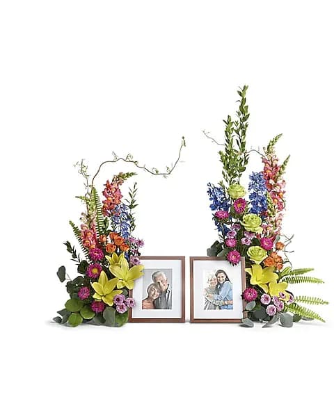 Loving Farewell Photo Tribute Bouquet - An awe-inspiring accent for your beloved photos at the service, this lush rose, lily and delphinium bouquet reflects a colorful life full of radiant memories.  Green roses, orange spray roses, yellow asiatic lilies, blue delphinium, orange snapdragons, hot pink matsumoto asters, and lavender button spray chrysanthemums are accented with curly willow, myrtle, sword fern, silver dollar eucalyptus, and lemon leaf. Arrangement does not include picture frame.