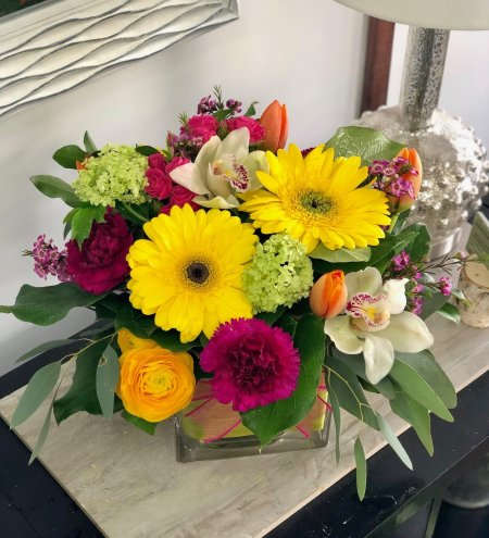 Lemon Lush Lemon Lush - A joyful burst of color and elegance, this low and lush arrangement features radiant yellow gerbera daisies paired with vibrant fuchsia carnations, soft white cymbidium orchids, and bright orange tulips. Accents of green hydrangea, golden ranunculus, and playful filler flowers are nestled among fresh eucalyptus and lush greenery. Arranged in a sleek glass cube vase, this design is perfect for bringing a touch of sunshine and sophistication to any space.