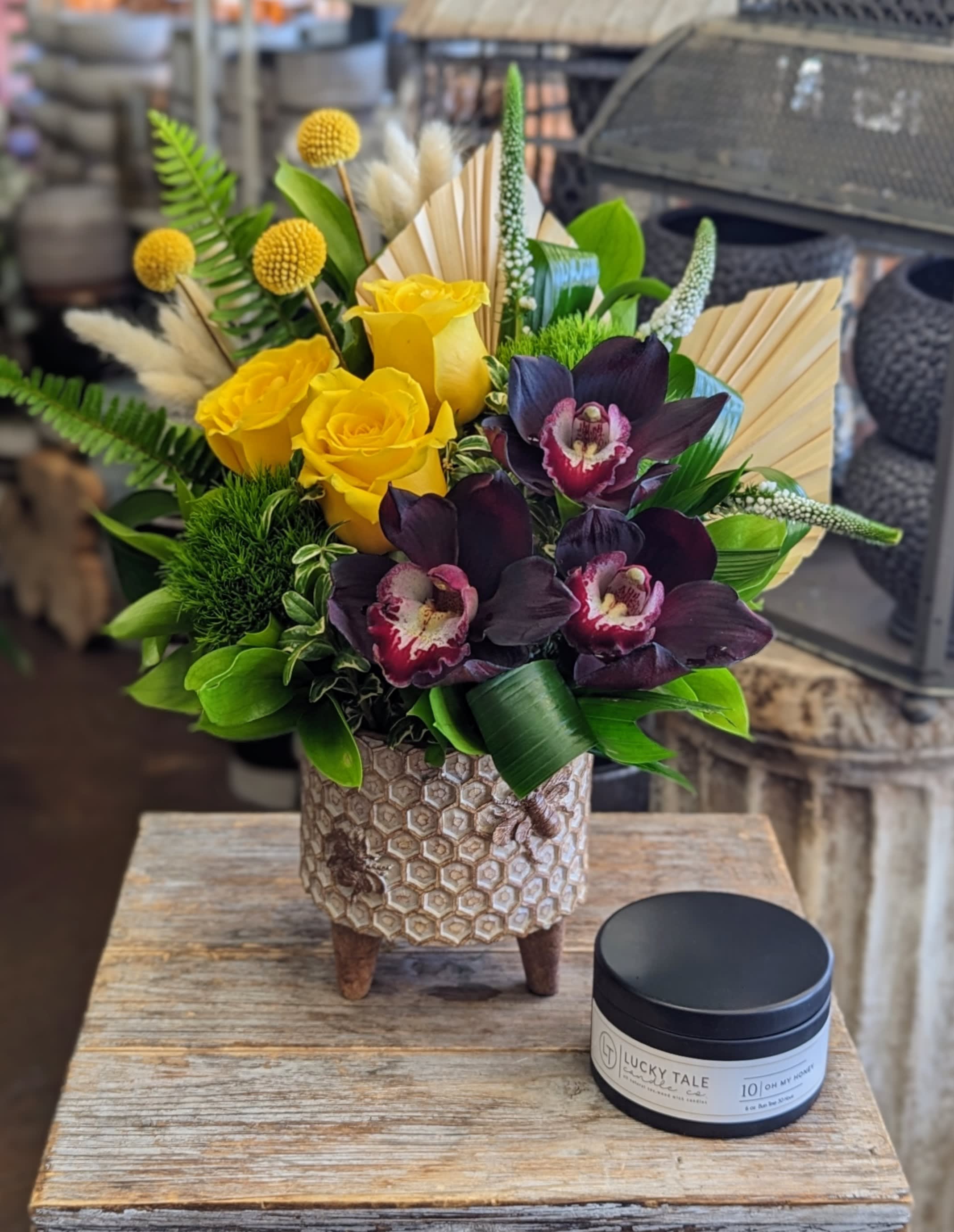 Le Miel - Includes A "Oh My Honey" Soy Candle Le Miel - Includes A "Oh My Honey" Soy Candle - Le Miel by Windmill Floral Expressions is a golden symphony of contrasts—where sunlight meets shadow, and bold design meets refined beauty. Nestled in a charming honeycomb-patterned ceramic planter perched on three modern legs, this floral arrangement glows with warmth, whimsy, and a quietly luxurious edge. At its heart, golden yellow roses shine like drops of liquid sunlight, while whimsical craspedia (billy balls) add sculptural delight with their spherical forms. In striking contrast, black cymbidium orchids bloom with velvety intensity—each petal deep and dramatic, encircling creamy centers kissed with magenta, like secrets spoken in candlelight. Delicate accents of cream bunny tails, dried palm fans, and green trick dianthus bring playful texture, while white veronica, ferns, and lush foliage add graceful movement and layers of fresh vitality. This radiant composition is paired with a Lucky Tale "Oh My Honey" soy candle—a sweet, warm scent that evokes golden afternoons and cozy serenity. Together, they make an unforgettable gift set that speaks to both the senses and the soul. Le Miel is perfect for birthdays, thank-yous, housewarmings, or as a statement of joy and elegance. Available for same-day flower and gift delivery in Las Vegas and Henderson.